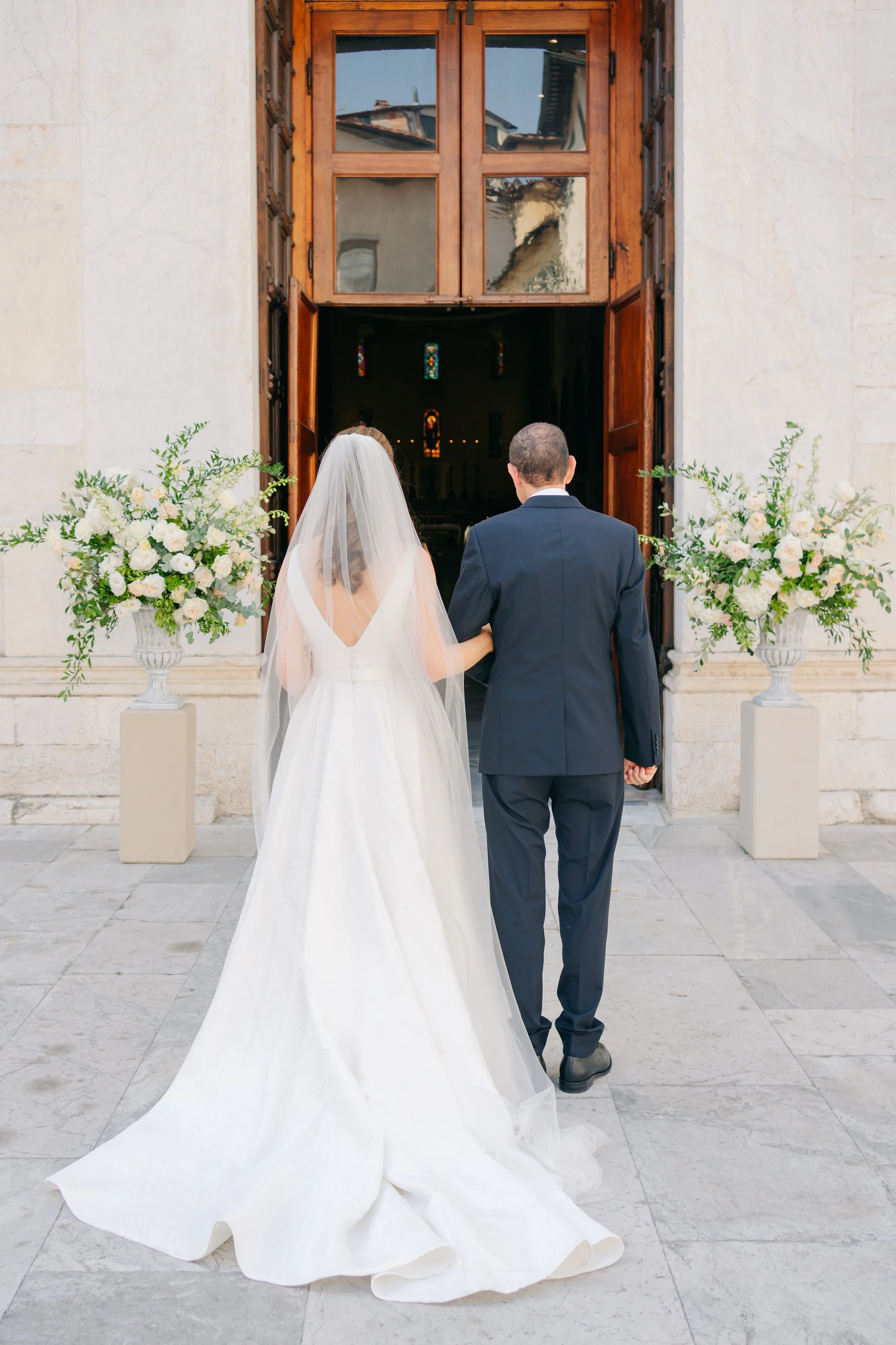 Sposa e sposo che entrano in chiesa per il matrimonio, vasi di fiori bianchi ai lati della porta, scena all'aperto con pavimentazione in pietra.