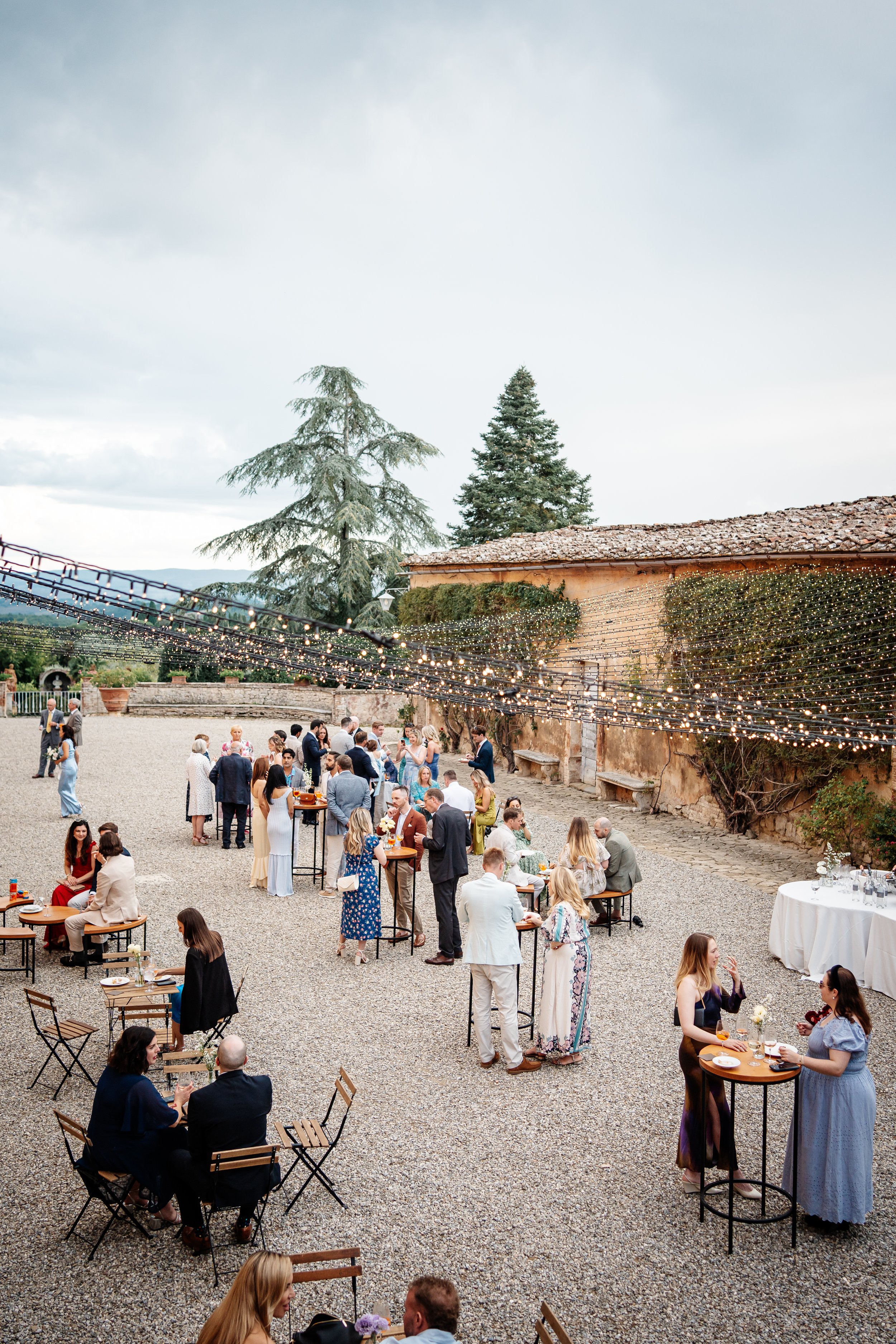 Una scena di festa all'aperto con molte persone che socializzano sotto luci decorative, su un'area di ghiaia con alcune sedie e tavoli, circondata da pareti di pietra e alberi al tramonto.
