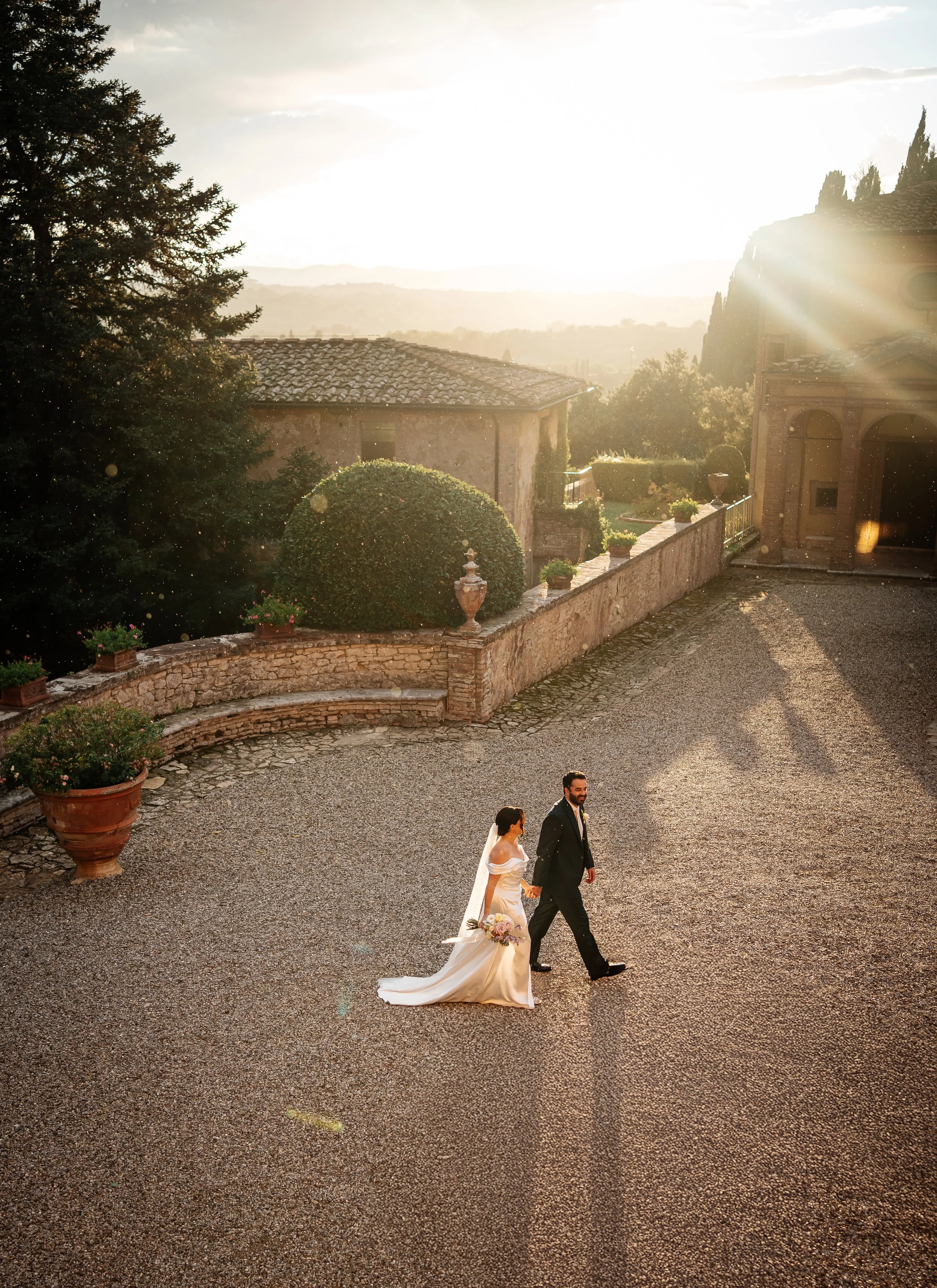 Un matrimonio cammina mano nella mano in un cortile al tramonto, con la sposa che indossa un abito bianco e il marito un abito nero.