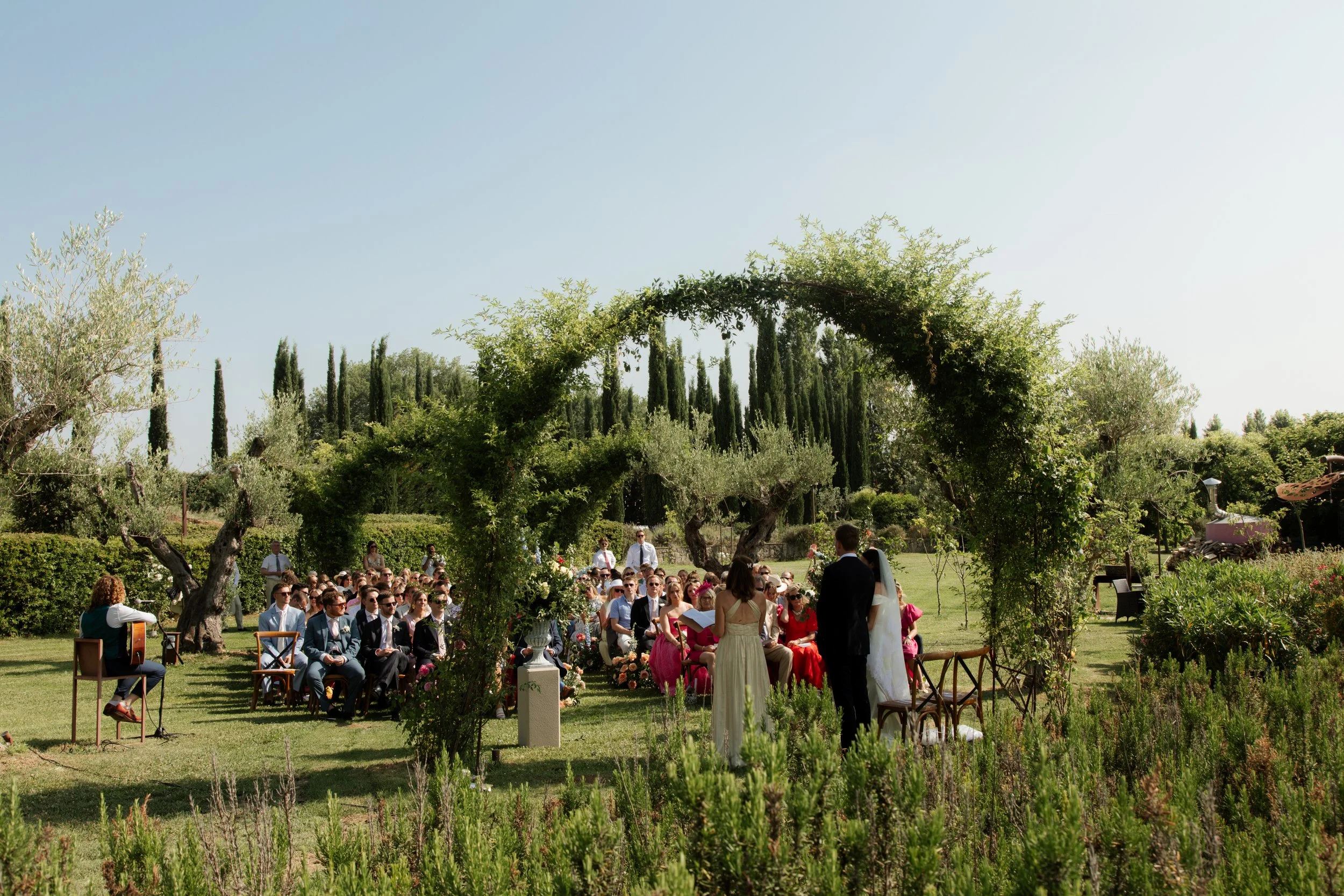 Cerimonia di matrimonio all'aperto sotto un arco di piante verdi, con sposi e invita presenti in un giardino rigoglioso.