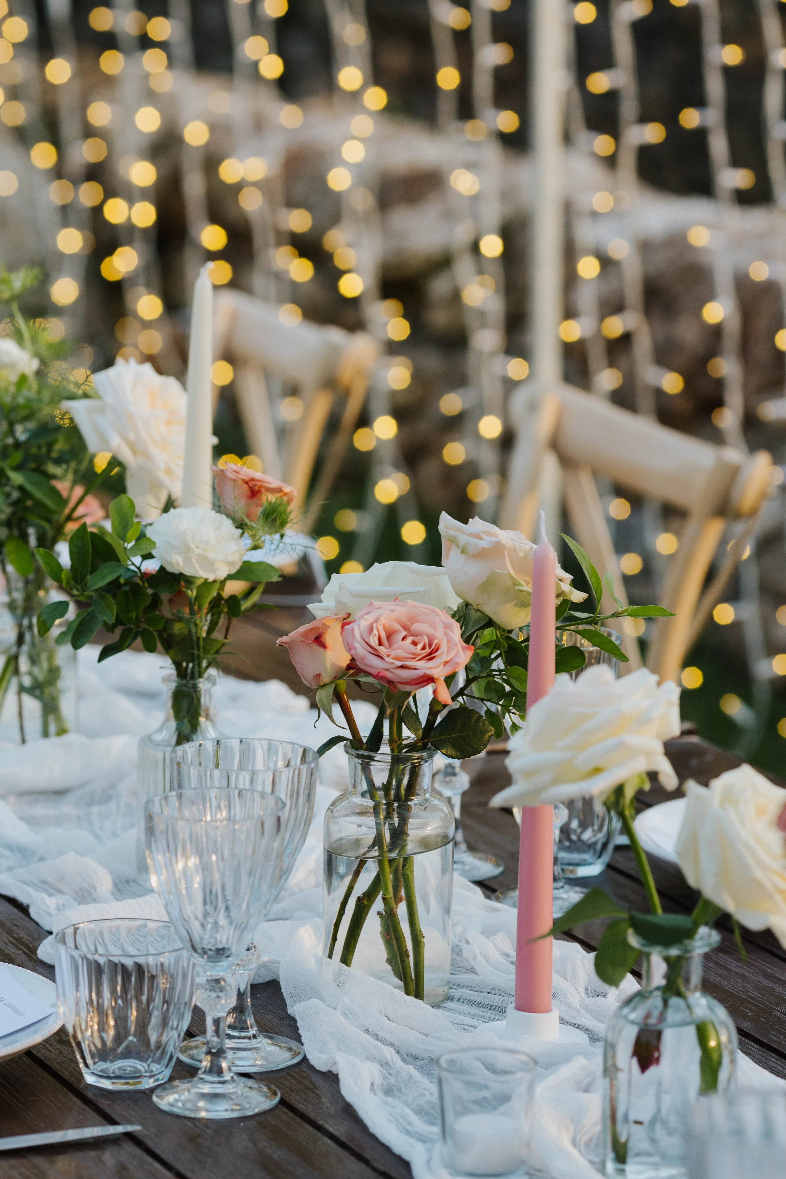 Tavola imbandita con vasi di fiori rosa e bianchi, candele rosa, bicchieri di vetro, tovaglia di pizzo bianco, sedie di legno e luci decorative gialle in sfondo.