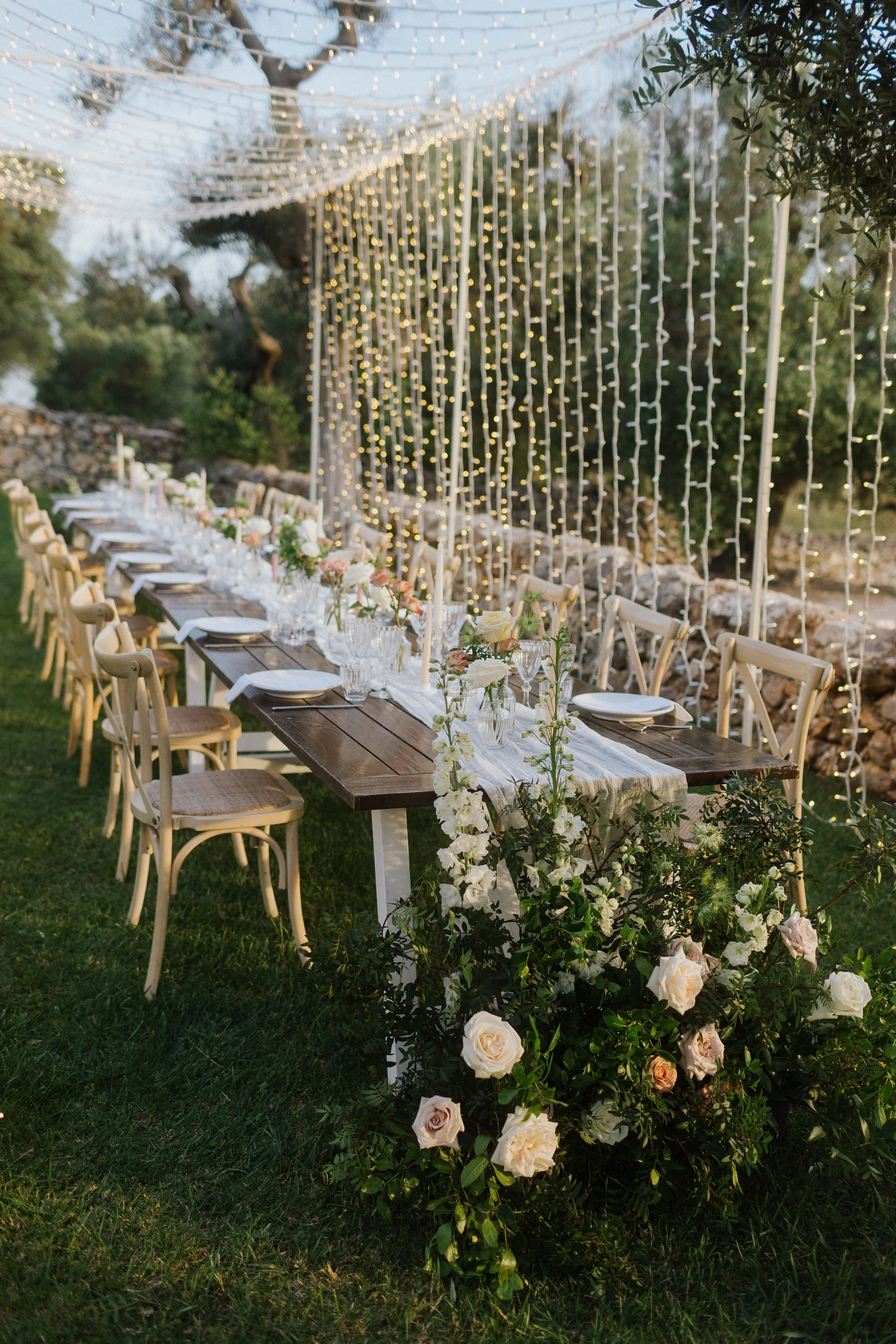 Tavolo all'aperto decorato con fiori e luci di festa, con sedie in legno e un ambiente naturale.