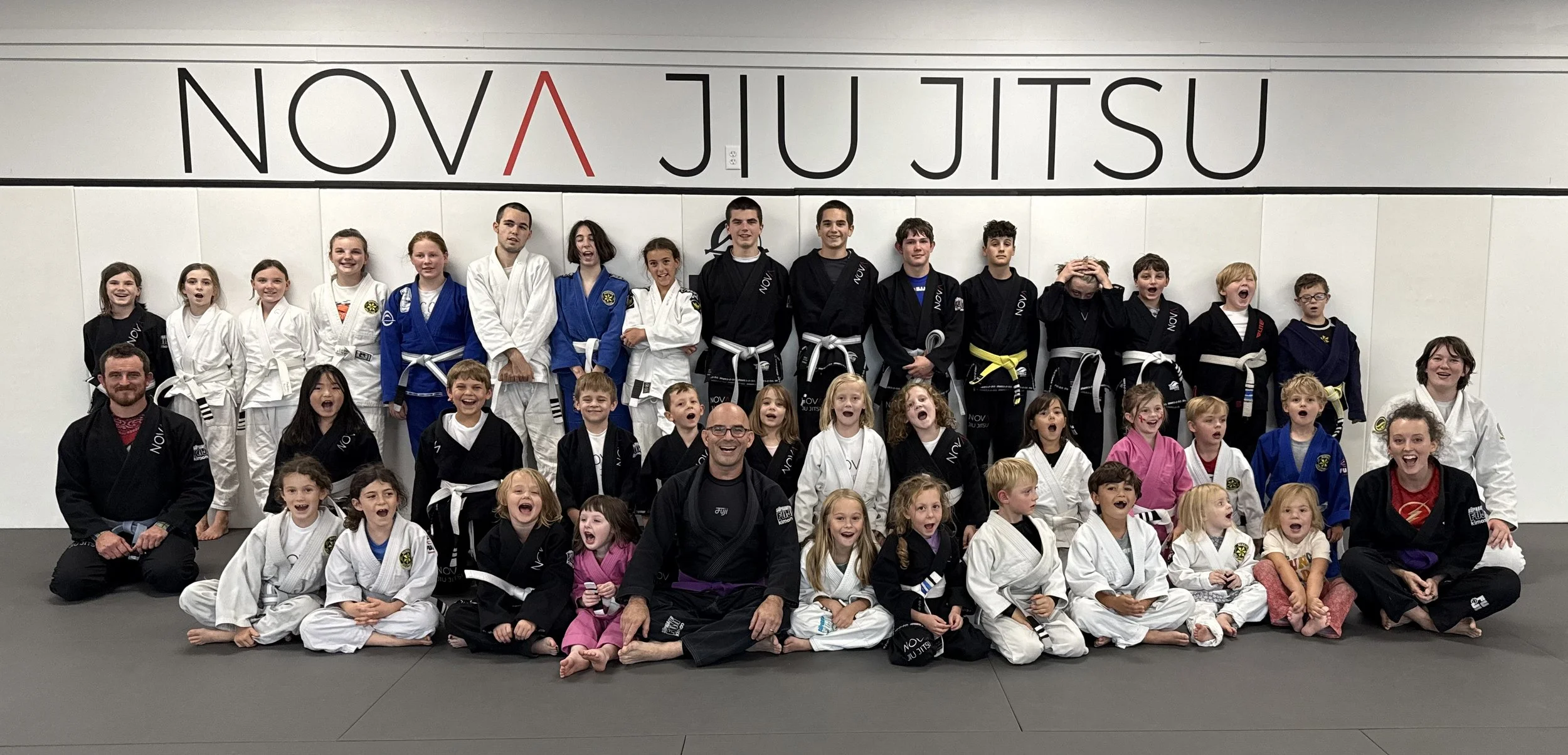 Group of children and instructors at a Brazilian Jiu Jitsu training facility, wearing gi uniforms and posing for a group photo in front of a wall with the words 'Nova Jiu Jitsu' displayed.