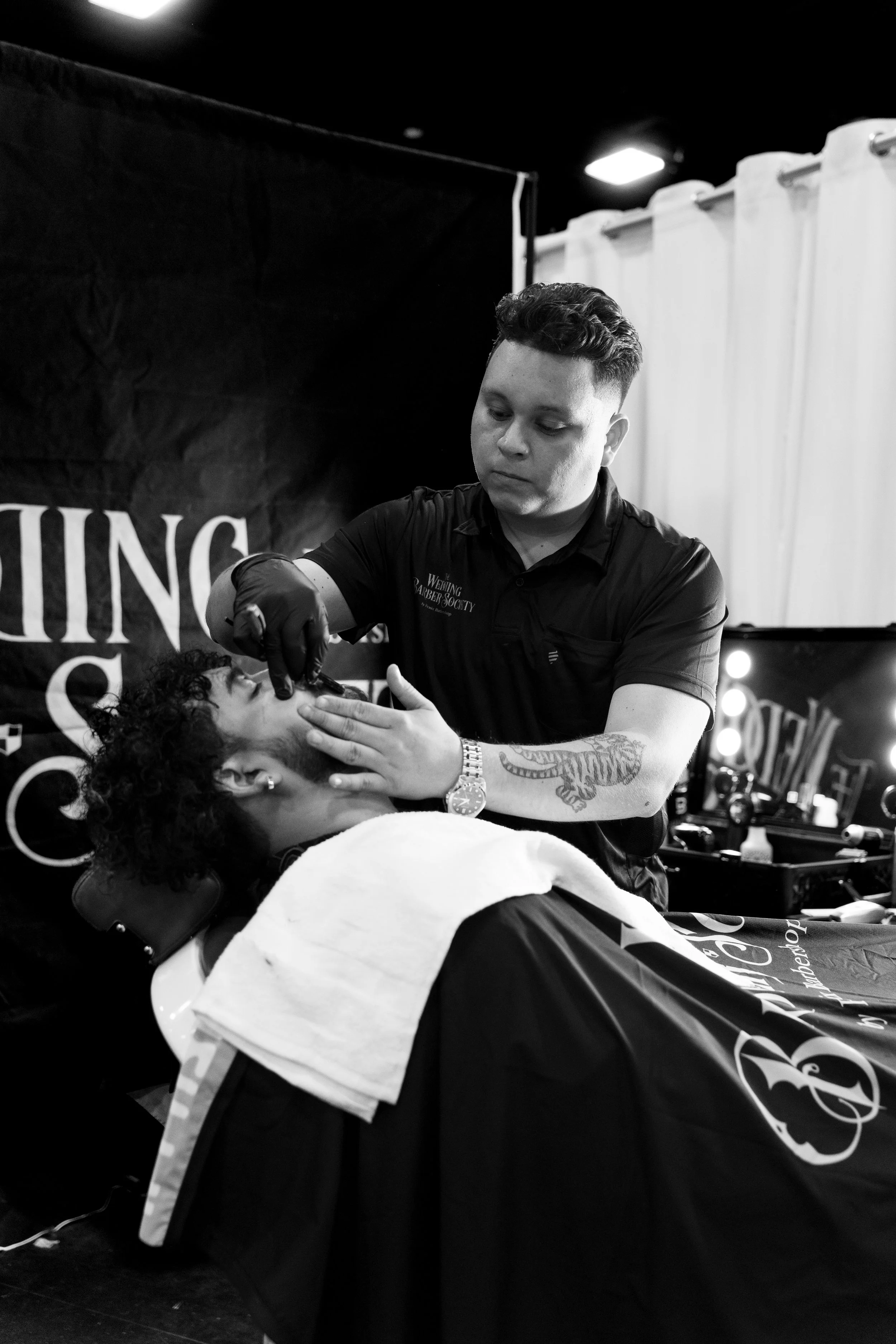 A man with a beard getting a haircut from a barber using a red comb and scissors, with another man, wearing a red baseball cap, observing in the background.