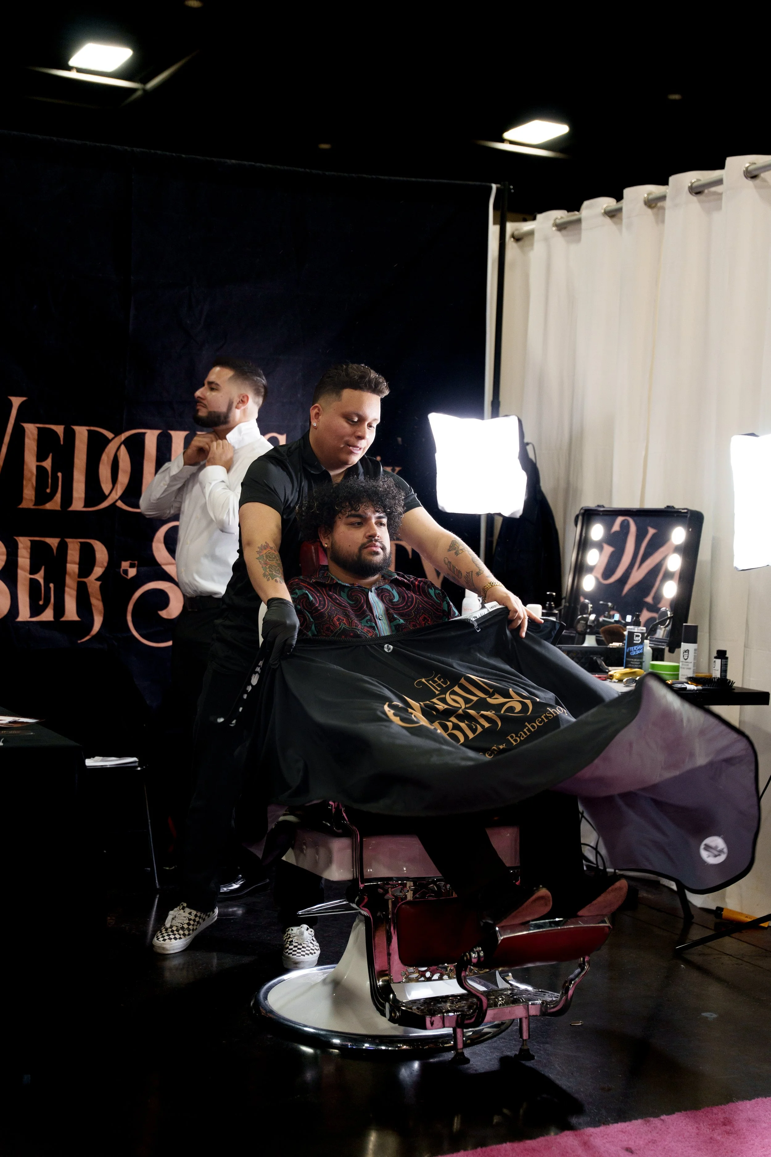 A young man with a fresh hairstyle sitting in a modern barber shop with neon lighting and graffiti art on the walls, wearing a barber cape.