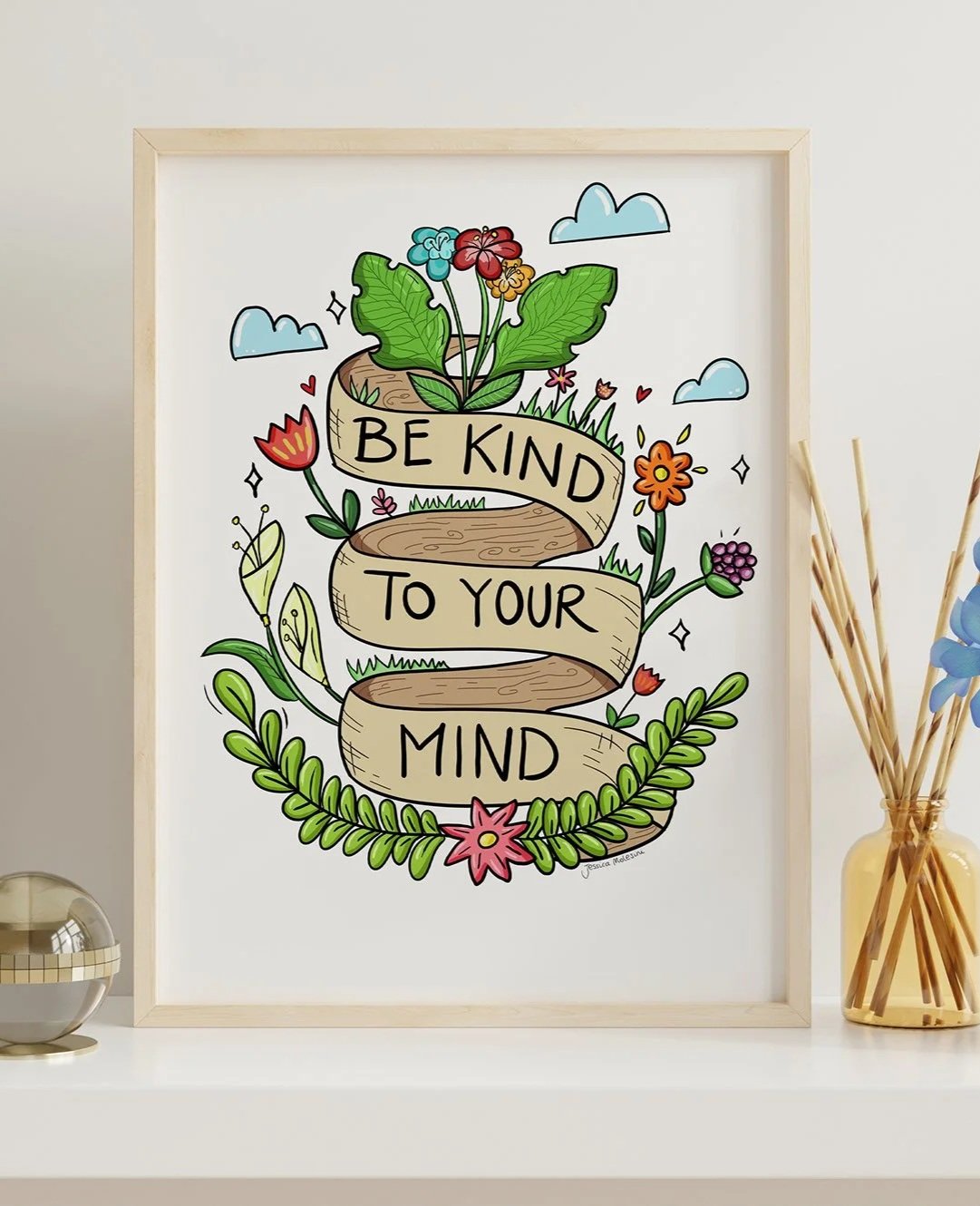 Decorative framed artwork with a floral design and a banner reading 'Be Kind To Your Mind' on a white surface, accompanied by a small metallic object and a glass jar with reed diffusers.