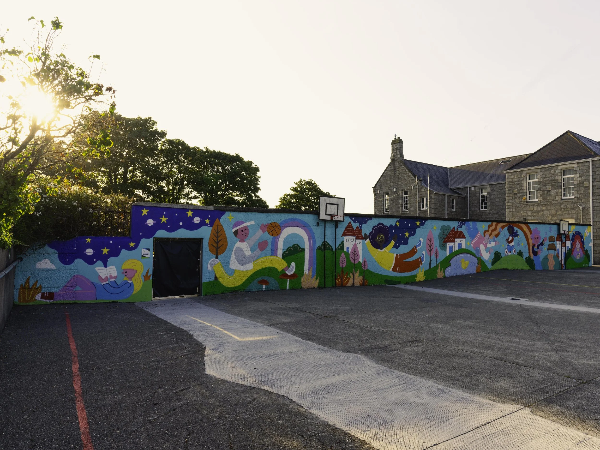 Colorful mural on a wall depicting children reading, playing, and engaging in various activities, with a blue sky, stars, trees, rainbows, houses, and other whimsical elements, adjacent to an empty parking lot and a stone building in the background.