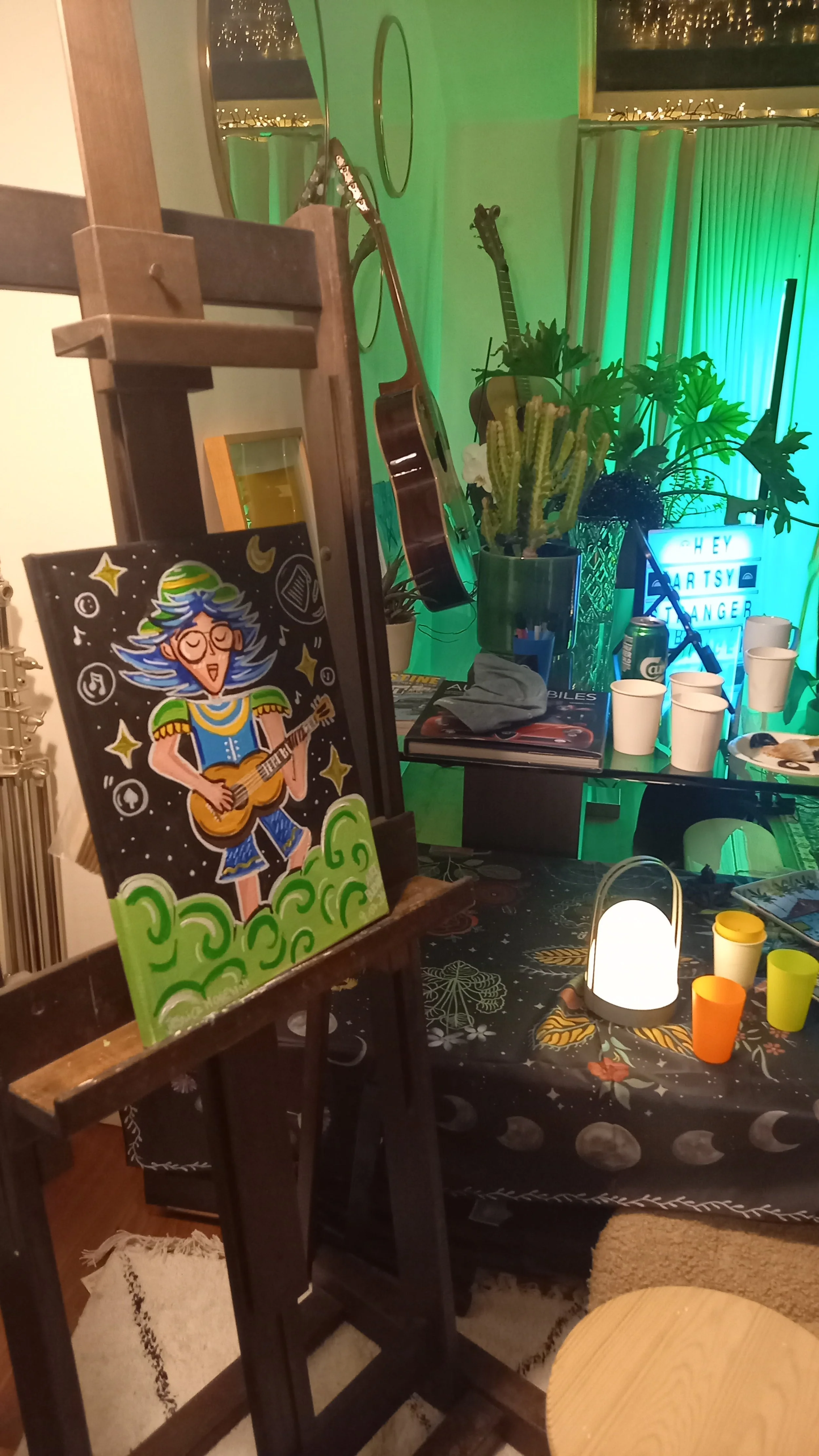 A decorated indoor space with a painting of a woman with blue hair playing a guitar, balloons with moons and stars, two guitars hanging on the wall, a table with cups, a small lantern, and various plants, including cacti, with a glowing sign in the b