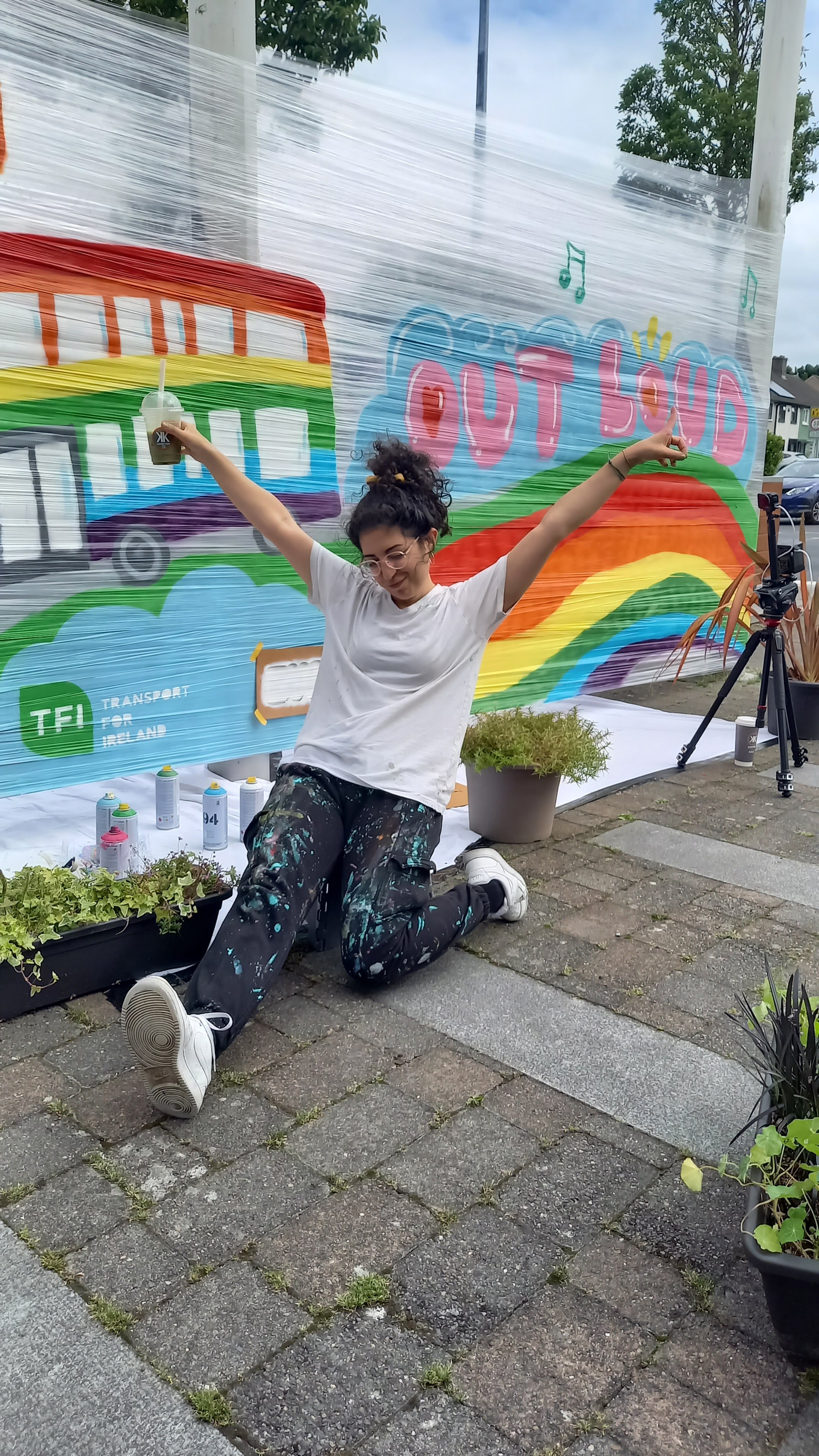 A young woman with curly hair tied up, wearing glasses and paint-splattered pants, poses on one knee in front of a colorful rainbow and cloud mural with the words 'Out Loud'. She is smiling, holding a drink in one hand and raising her arms in celebra
