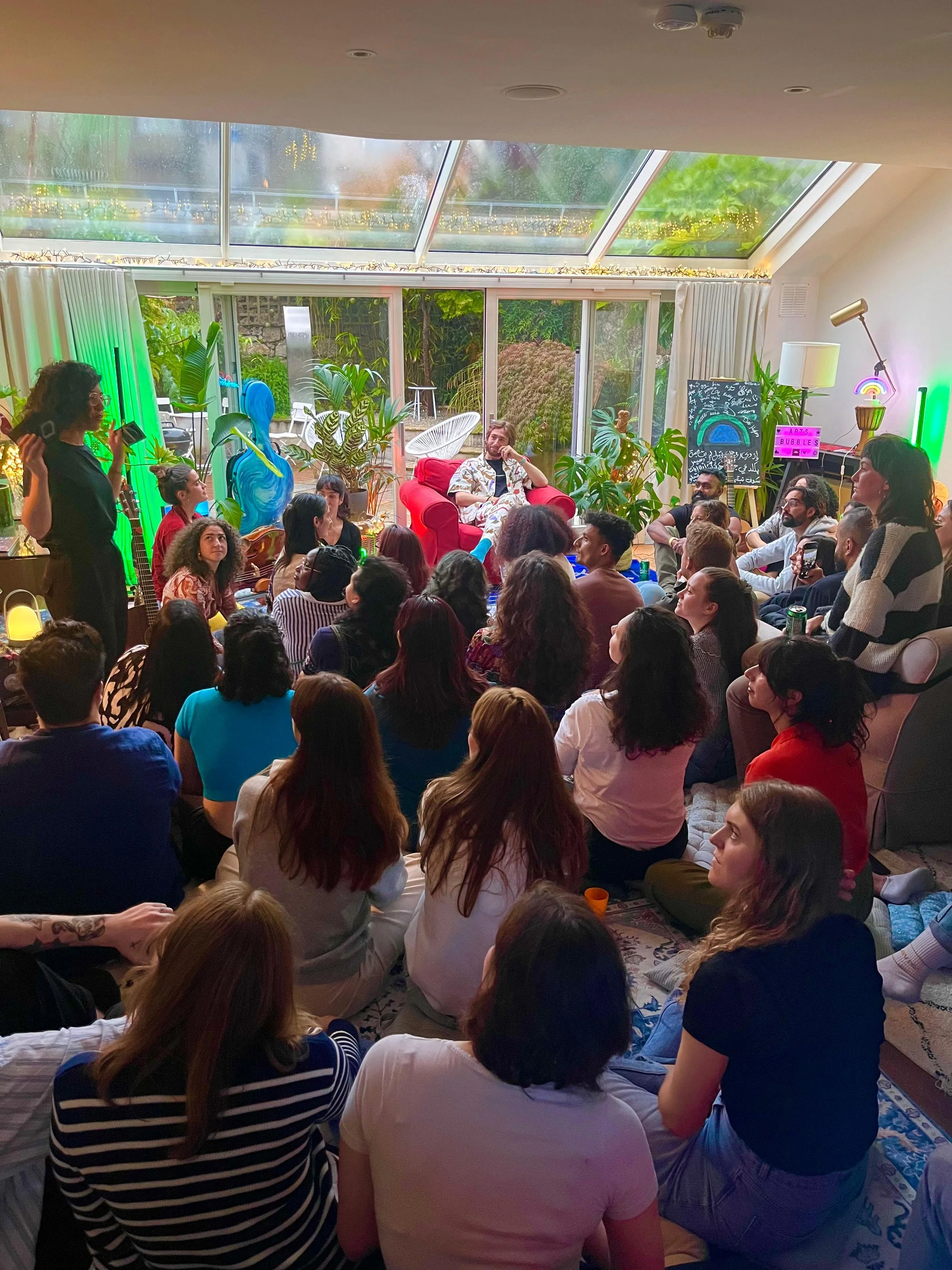 People gathered in a cozy living room, sitting on the floor and couches, watching a woman speak at an indoor event. The room has large windows with a garden view, colorful plants, and decorative art.
