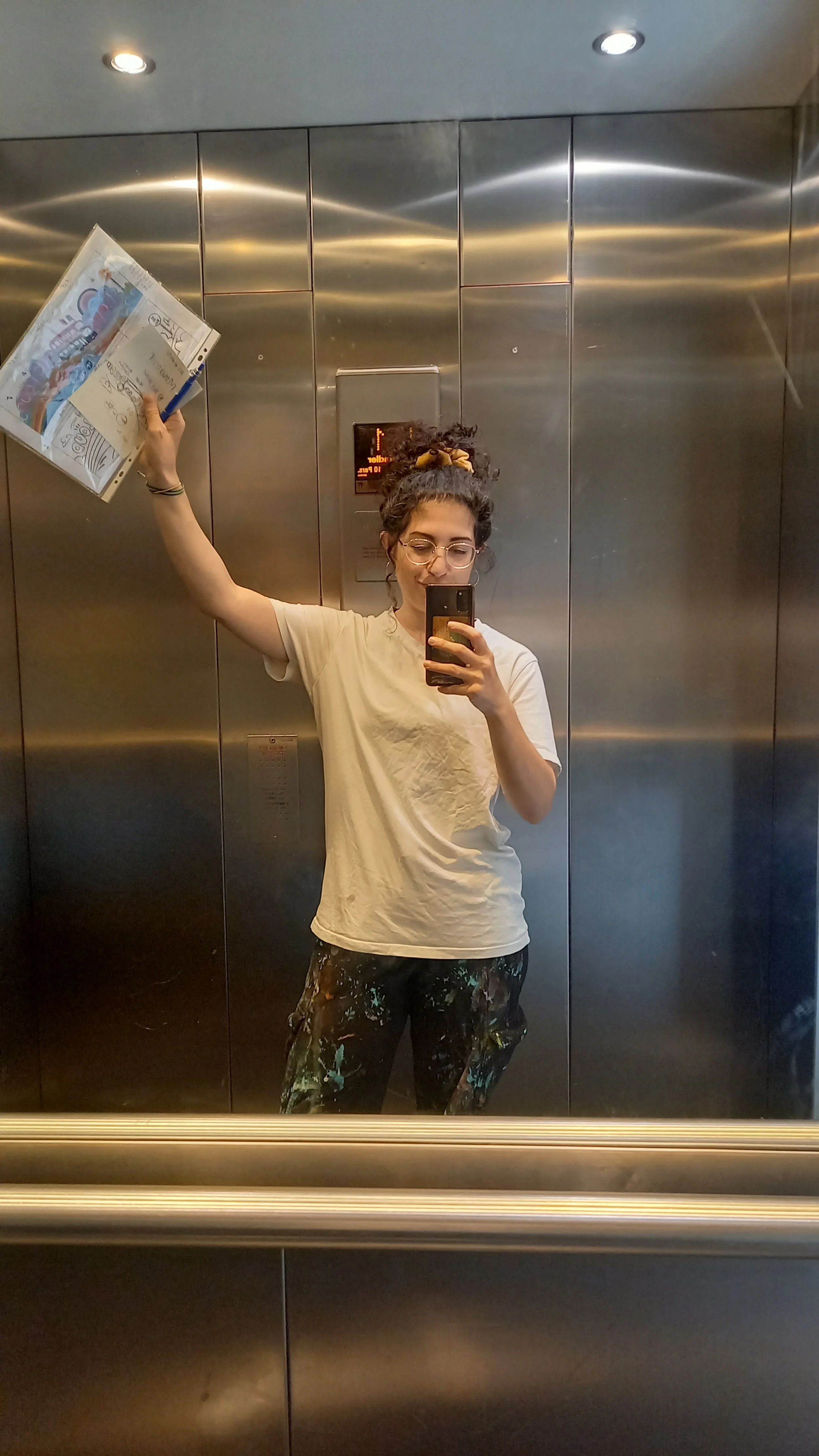 A woman taking a mirror selfie inside an elevator. She is holding a magazine in her left hand and smiling. She has curly hair tied up with a yellow scrunchie and is wearing glasses, a beige T-shirt, and dark pants with a colorful paint splatter patte