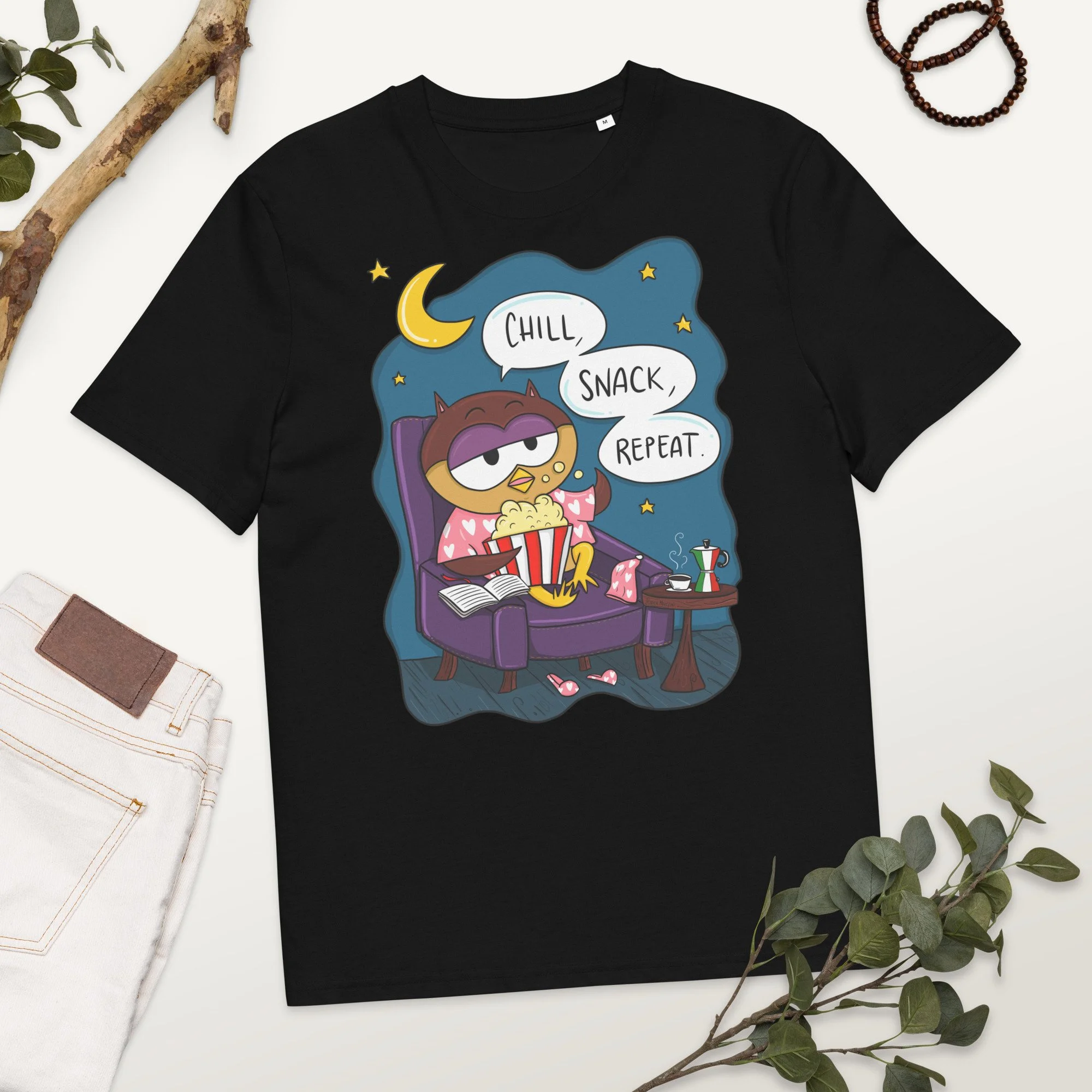 Black T-shirt with cartoon owl wearing pink pajamas sitting on a purple armchair, holding popcorn, with open book nearby, under a night sky with a crescent moon and stars. Speech bubble says, 'Chill, snack, repeat.'