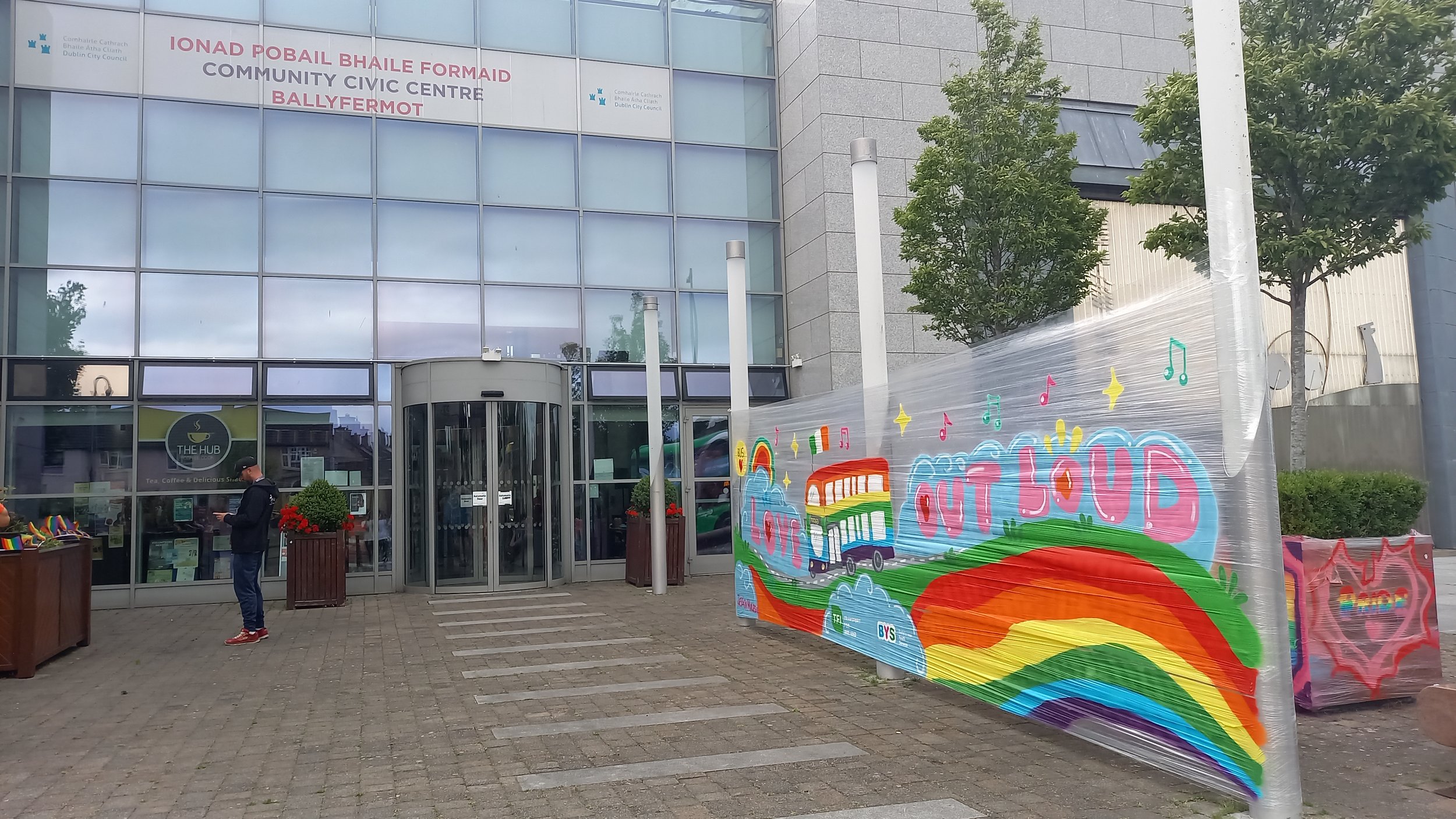 Entrance of a modern civic center with glass doors, two potted plants, and a rainbow-colored banner displaying rainbow arcade game graphics and the words "OUTLOUD".