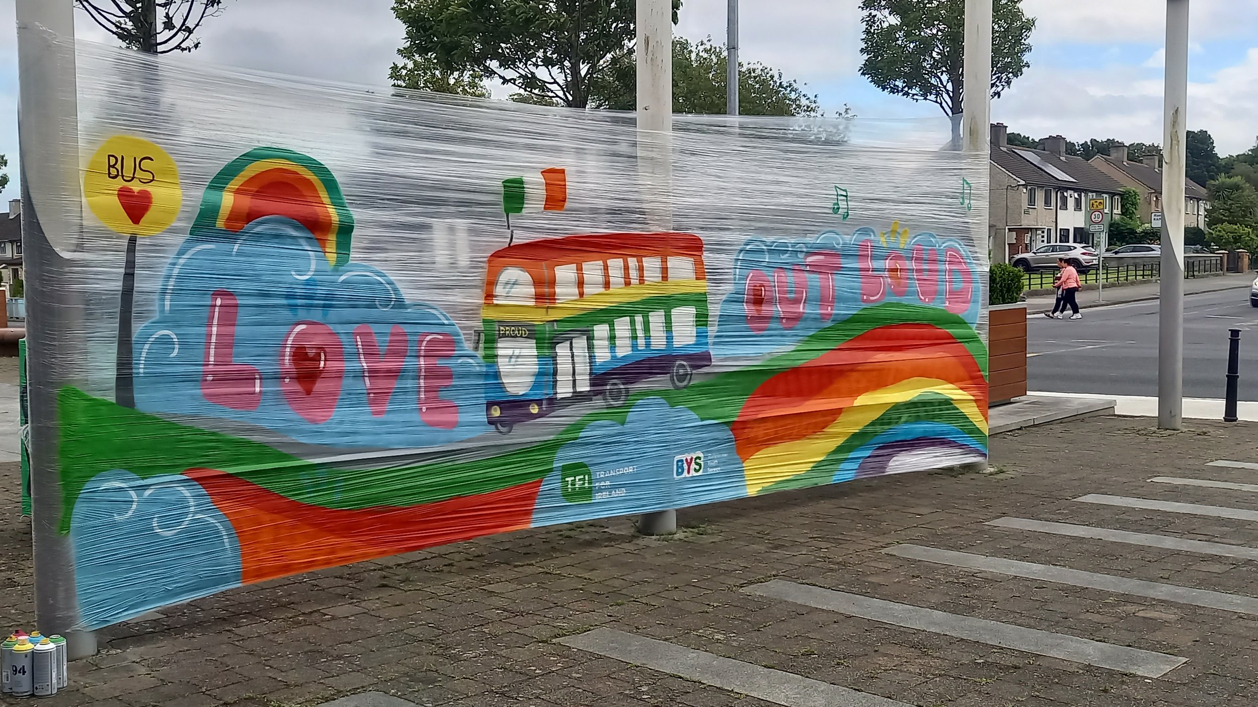 Colorful outdoor mural with a rainbow, a rainbow bus, and the words 'LOVE OUT LOUD' on clouds, with a landscape of rolling hills and clouds.