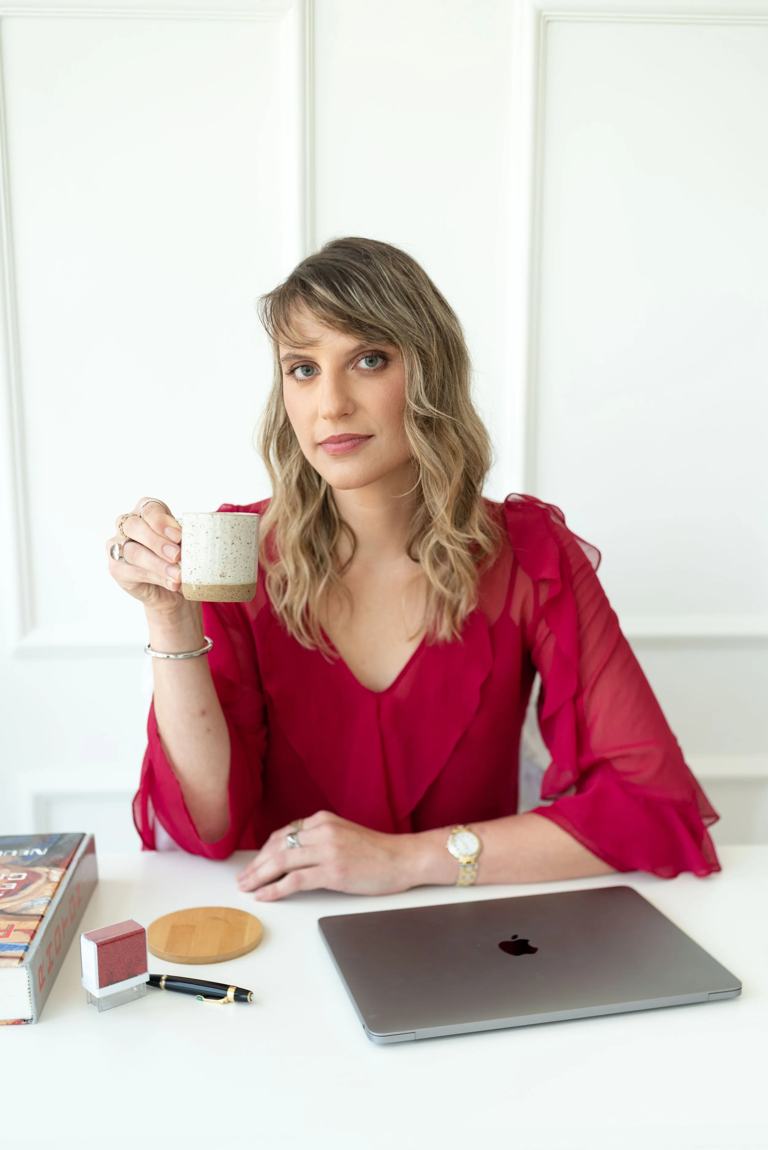 Mulher de cabelos castanhos, vestindo blusa vermelha, segurando uma xícara, sentada em uma mesa com laptop e objetos de escritório.