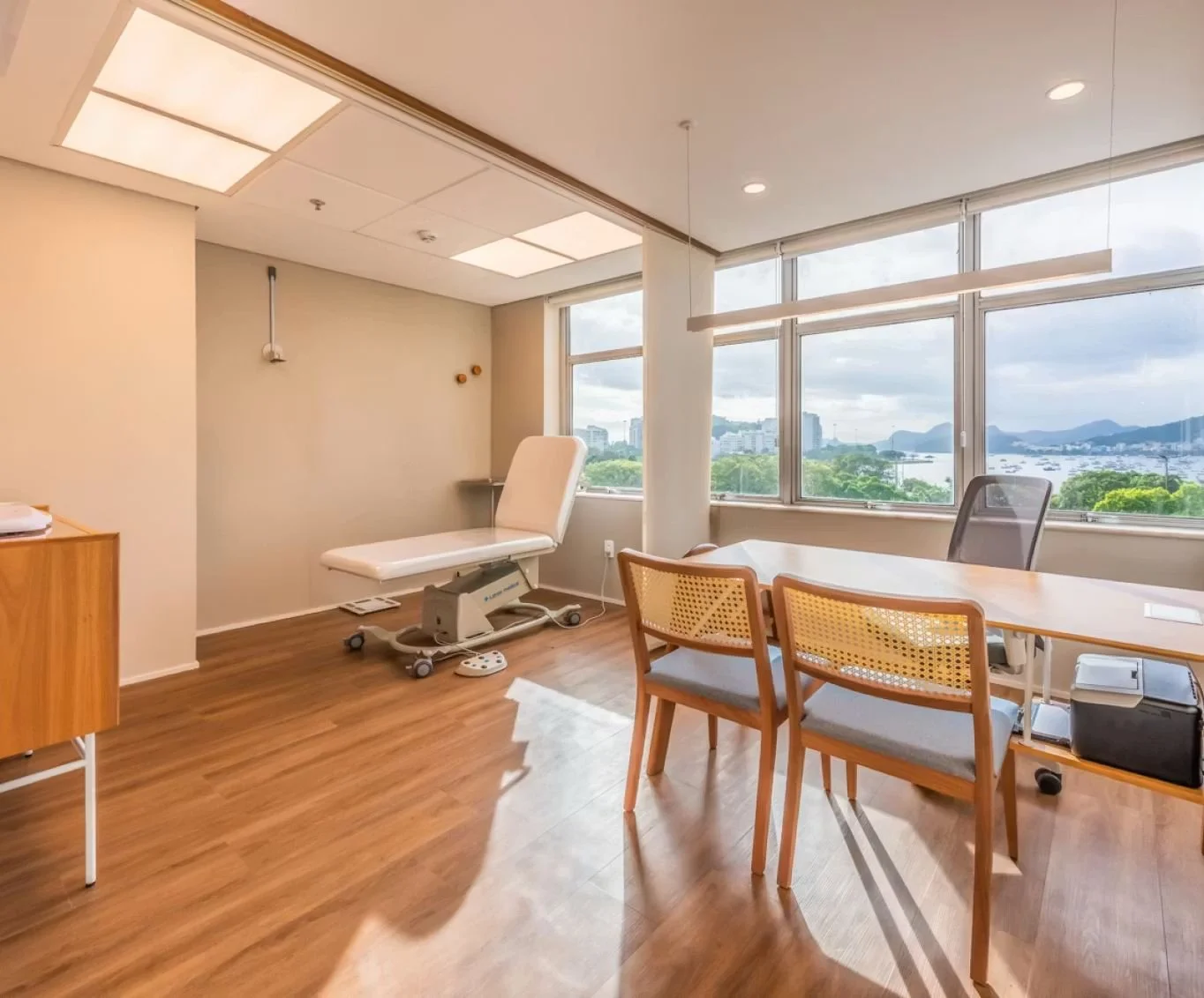 Consultório médico com mesa de exame, cadeira de escritório, mesa de madeira, vista para a cidade e o mar através de janelas grandes