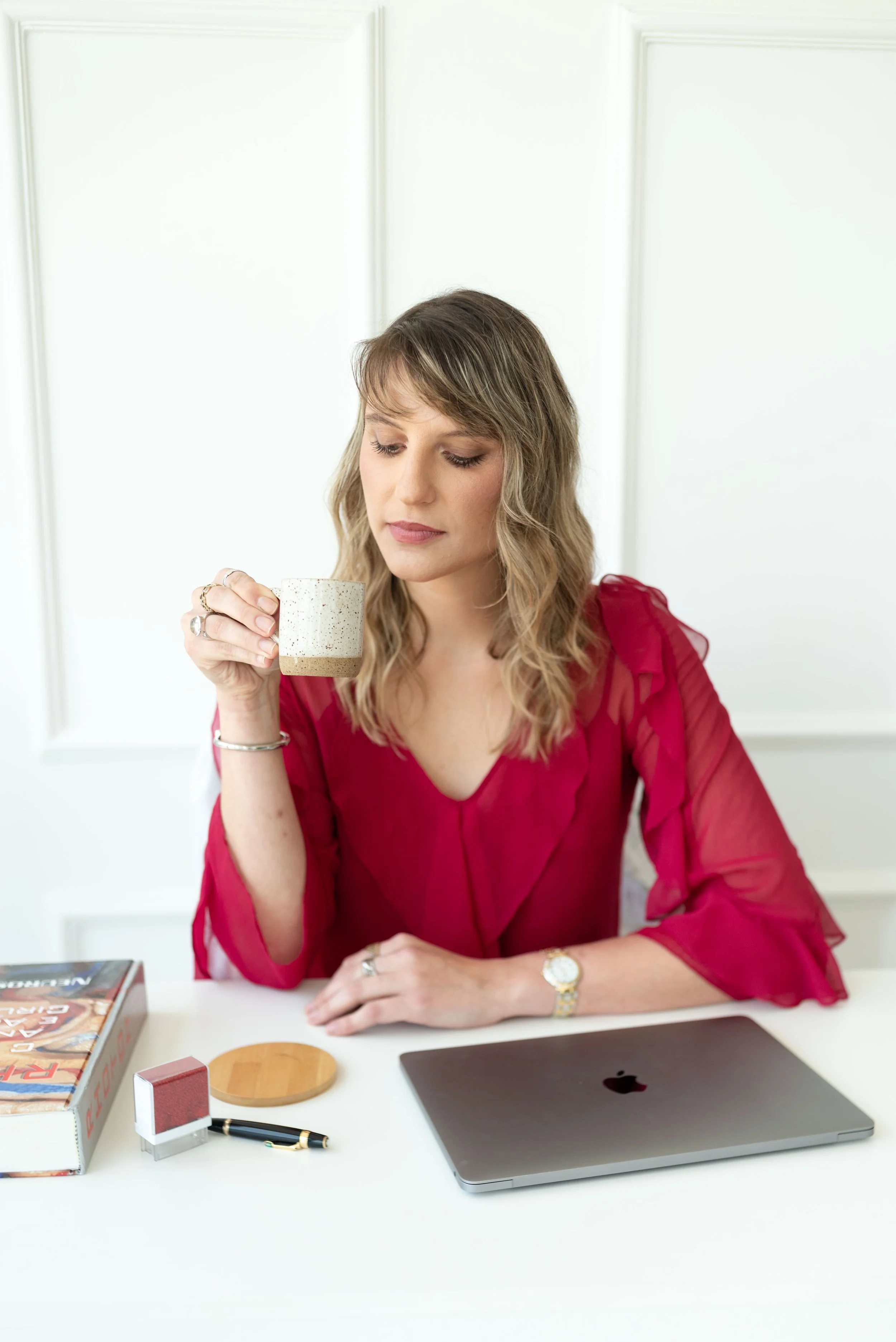 Mulher sentada à mesa segurando uma xícara de café, roupa vermelha, ao lado de laptop, livro, caneta, bloco de notas e mesa redonda de madeira.