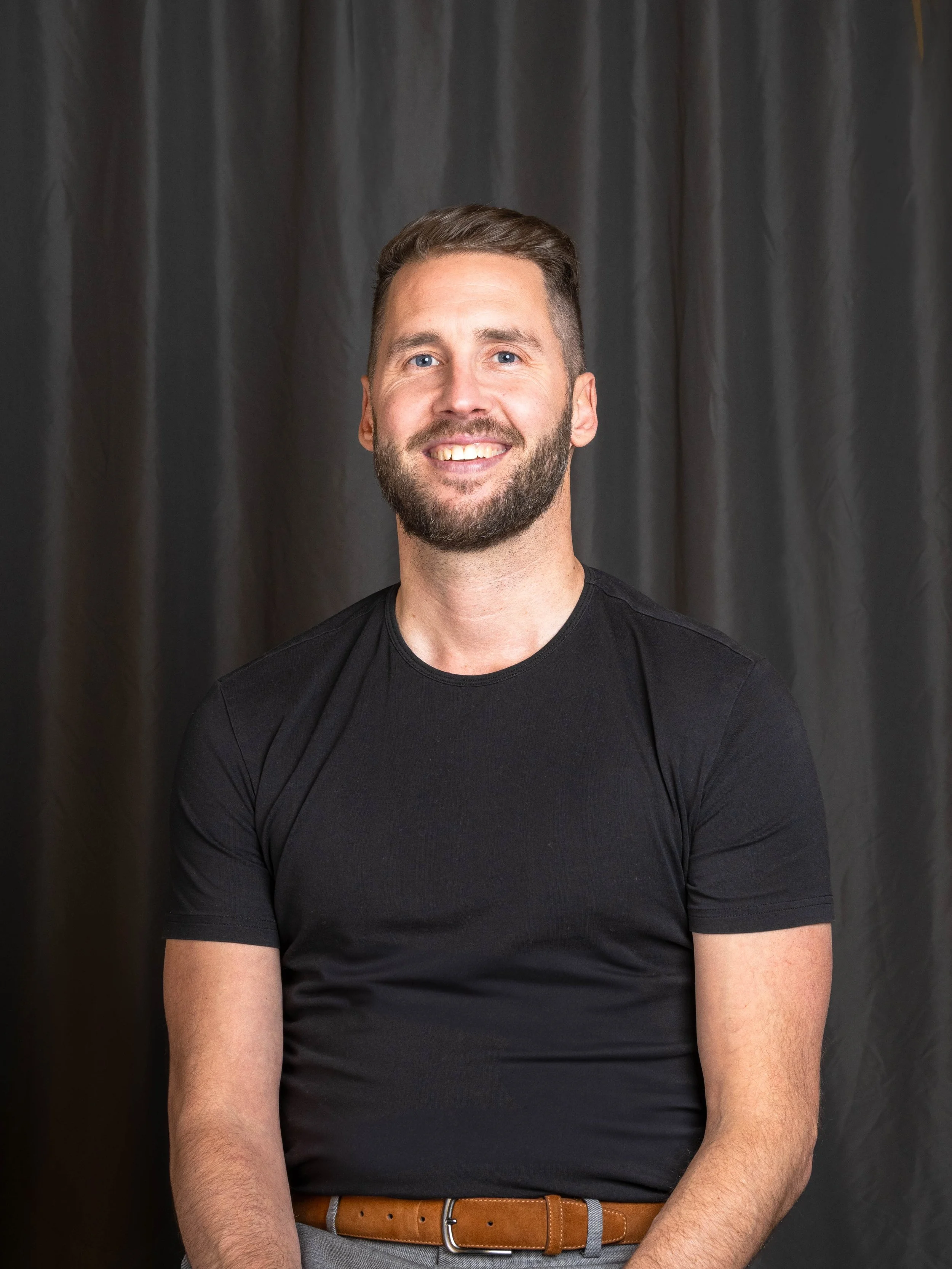 Portrait of a man with a beard and short hair, wearing a black t-shirt, smiling, with a dark curtain background.