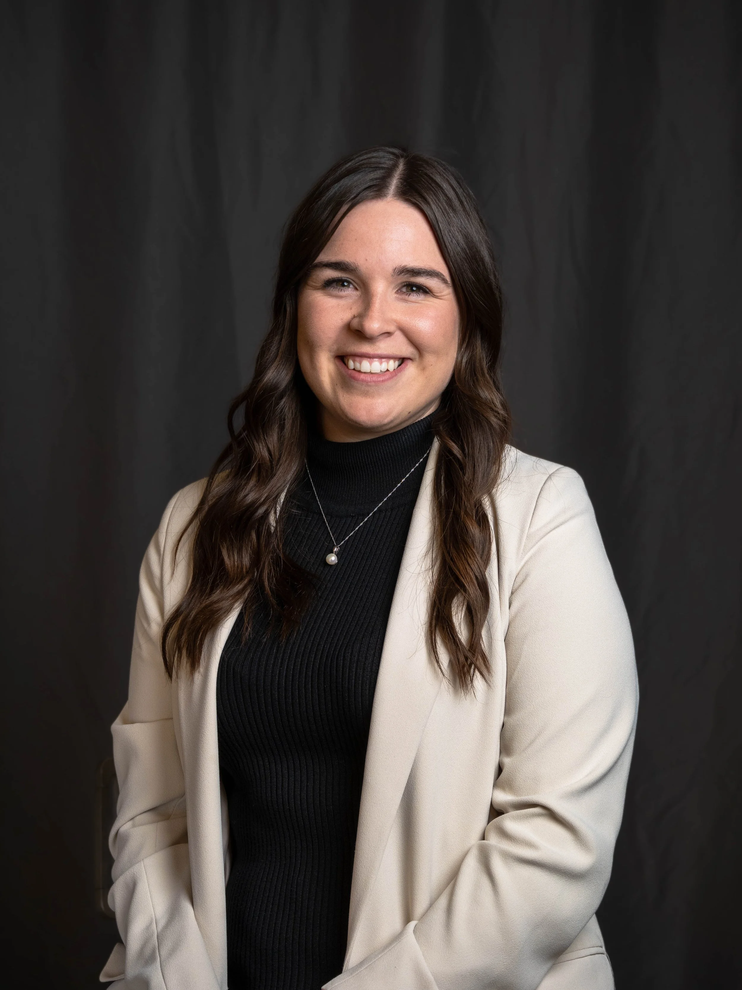 A young woman with long brown hair, smiling, wearing a cream blazer over a black turtleneck, and a silver necklace with a pearl pendant, against a dark gray background.