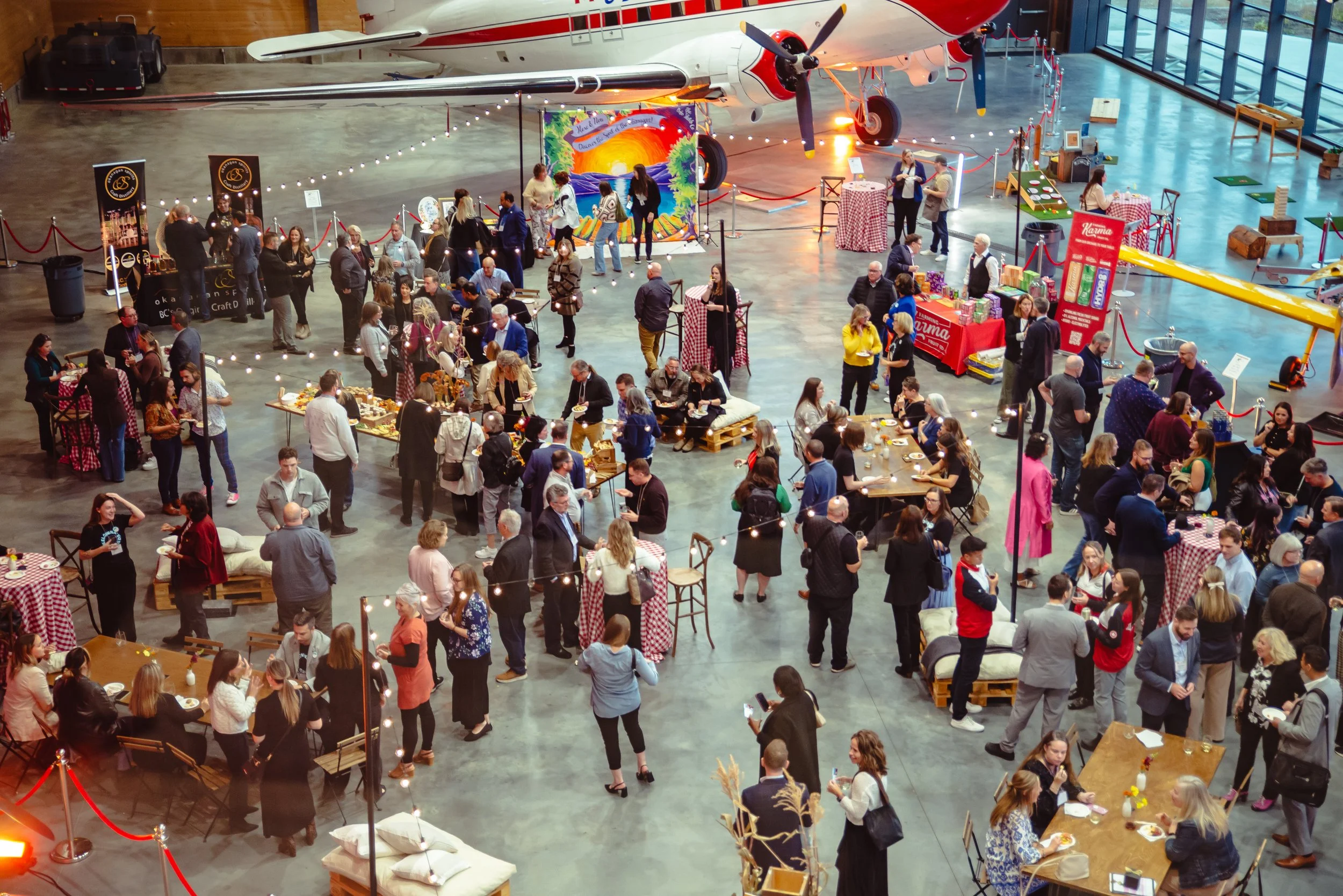 An indoor event at a museum or aircraft hangar with a large vintage airplane displayed overhead. People are gathered around dining tables with checkered tablecloths, enjoying food and drinks. There are various booths and informational displays, with some people standing and chatting, and others seated at tables. String lights hang above, creating a festive atmosphere.