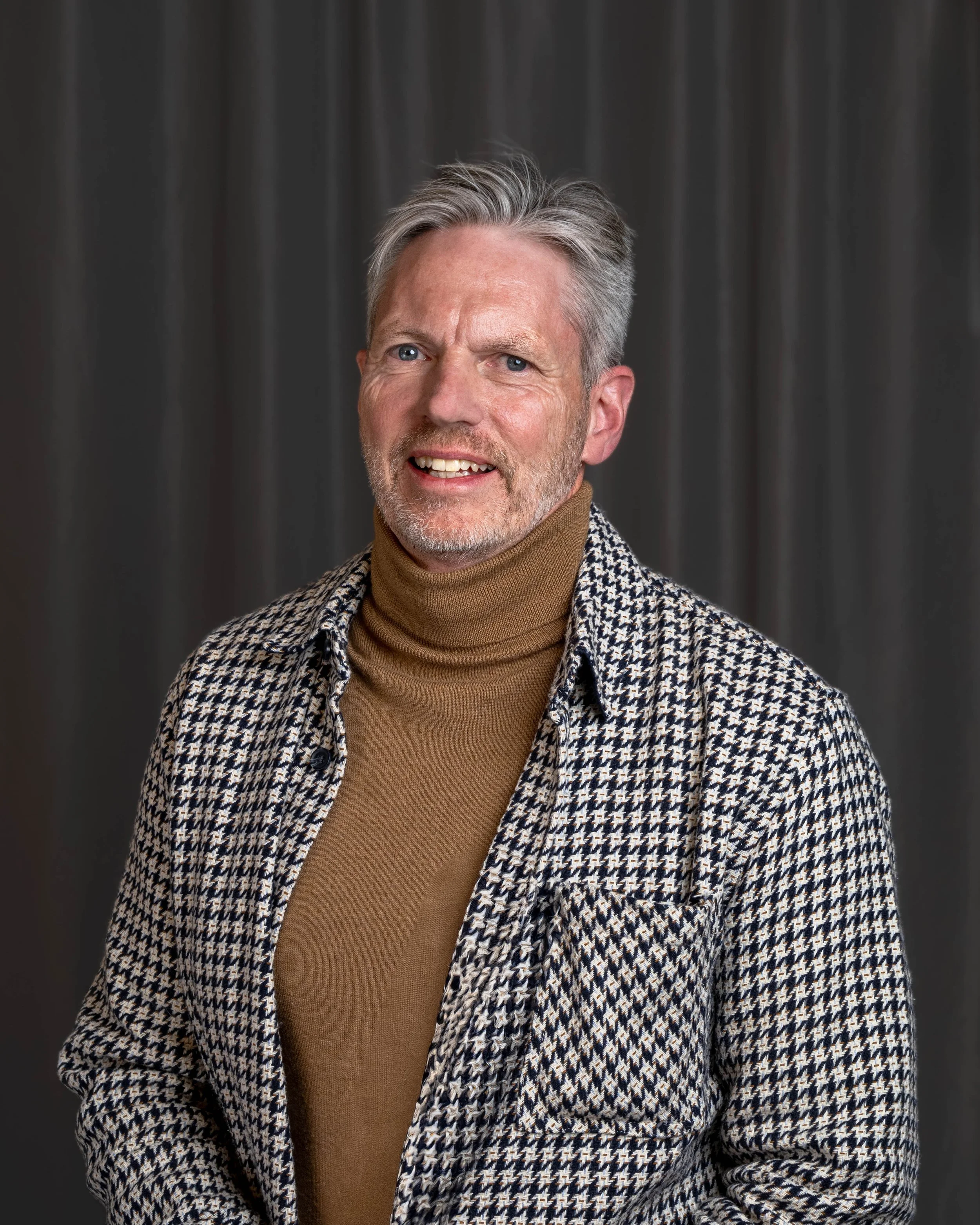 Portrait of a middle-aged man with gray hair and beard, wearing a houndstooth patterned blazer and brown turtleneck sweater, standing in front of a dark gray curtain.