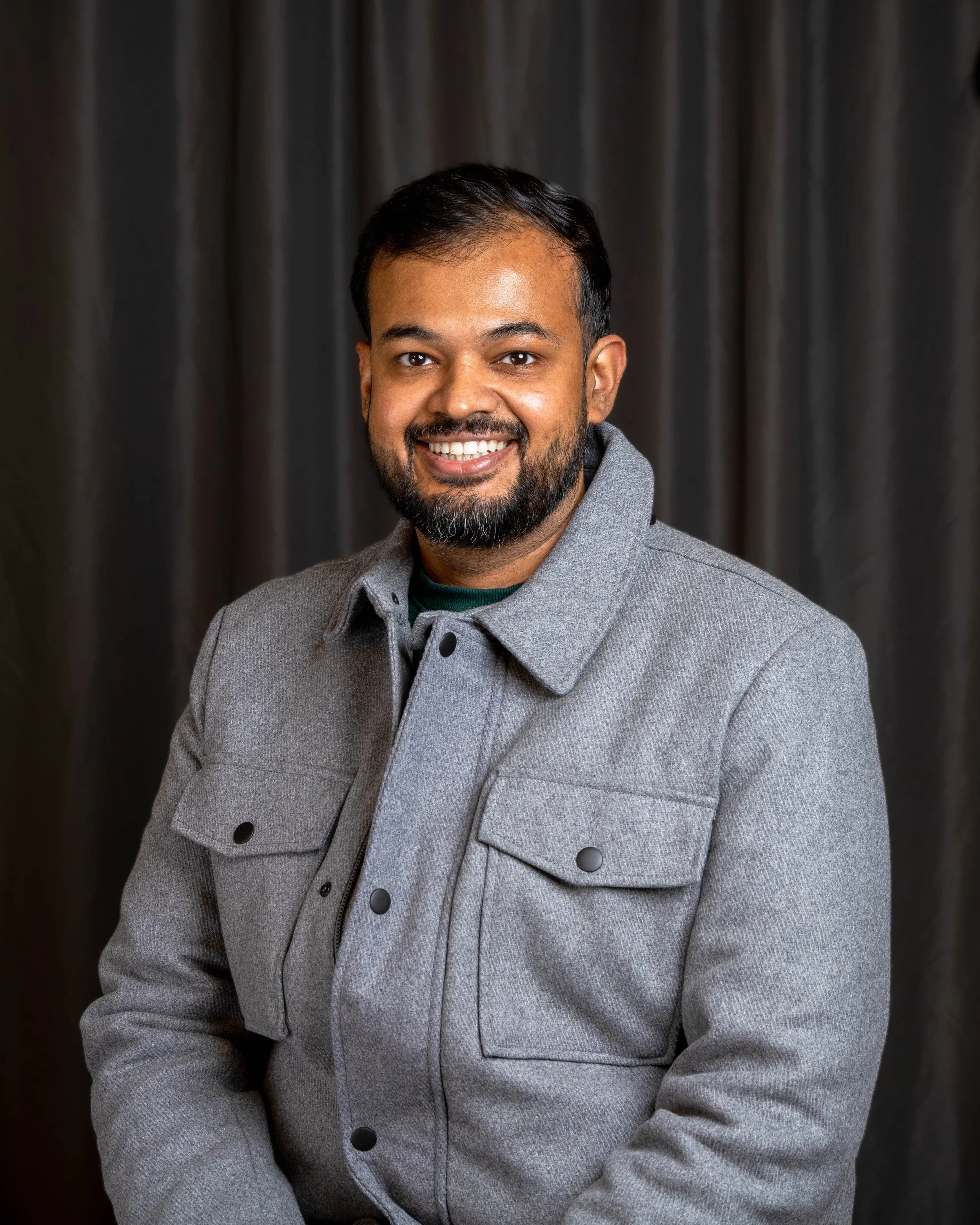 A smiling man with a beard, wearing a gray wool jacket, standing in front of a black curtain.