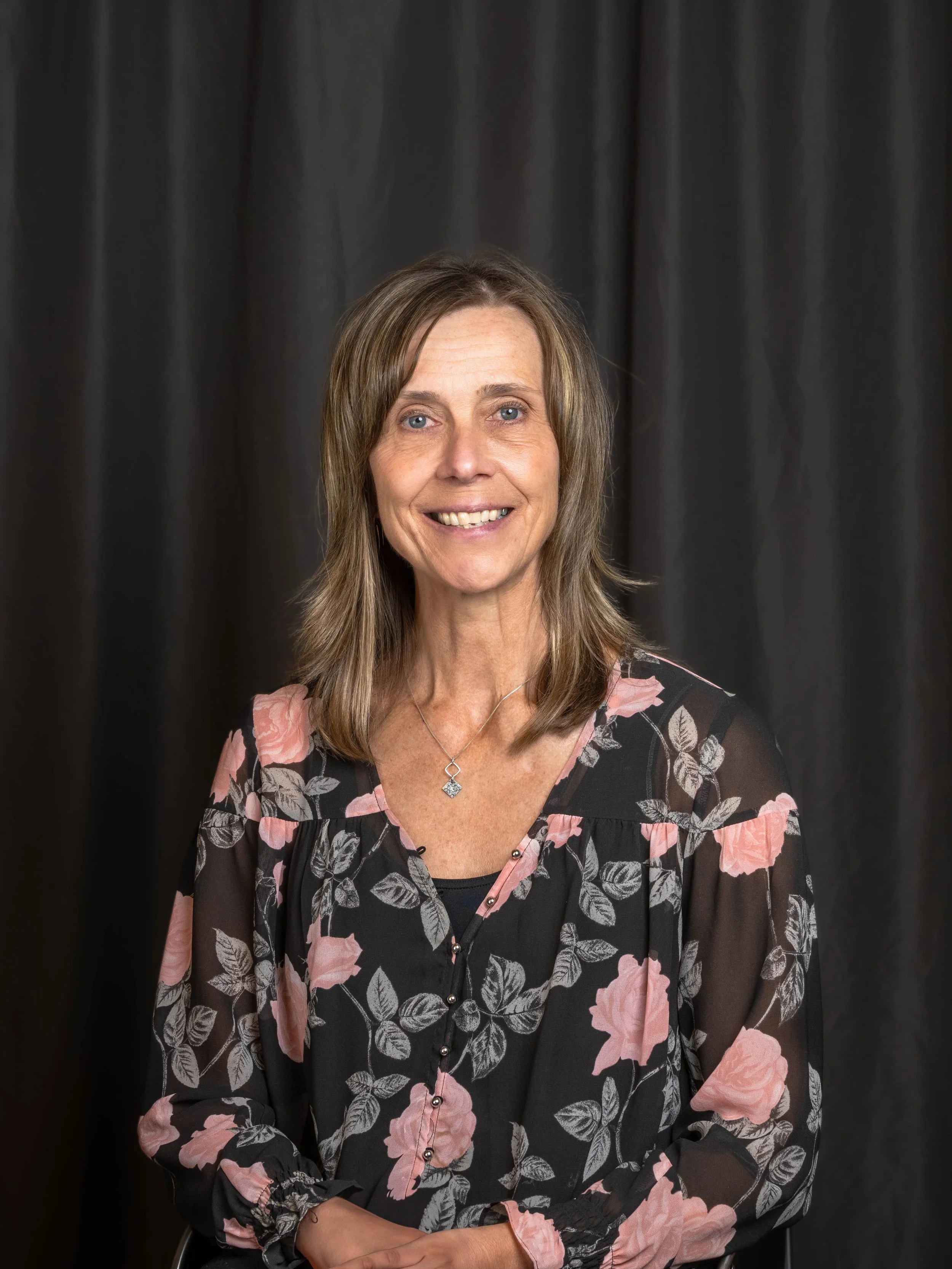 A smiling woman with shoulder-length brown hair wearing a black floral blouse with pink flowers, standing in front of a black curtain.