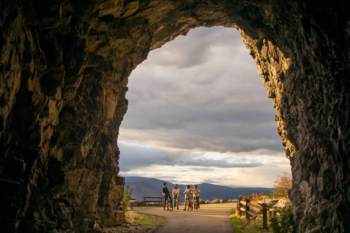 View from inside a large rock tunnel looking out at four people with bikes standing on a scenic overlook, mountains in the background, cloudy sky overhead.