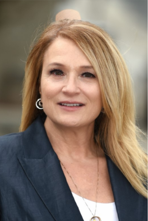 A woman with shoulder-length blonde hair, wearing a navy blazer, white top, and hoop earrings, smiling outdoors with blurred background.