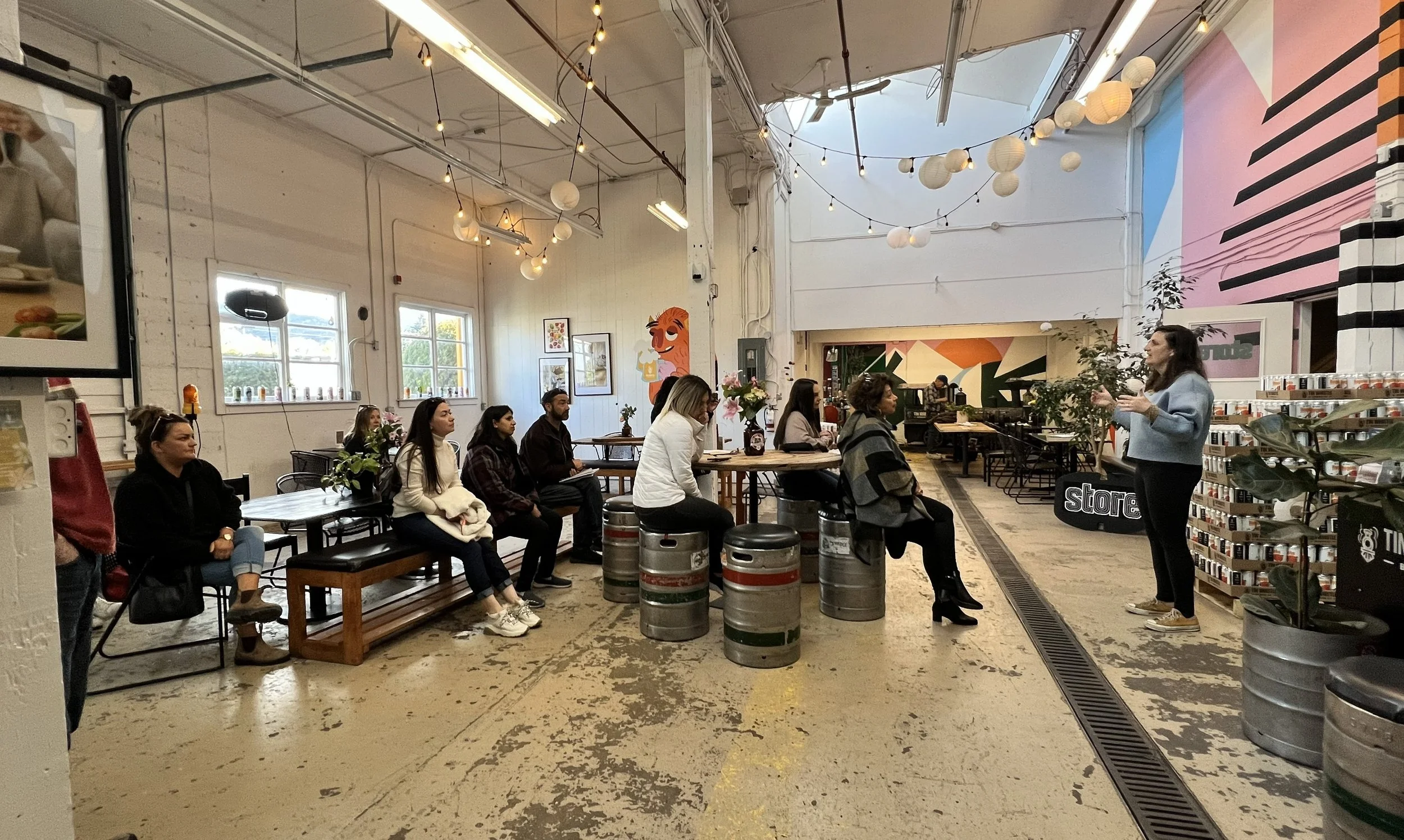 A woman giving a presentation to a seated audience inside a cafe or creative space with colorful wall art and string lights hanging from the ceiling.