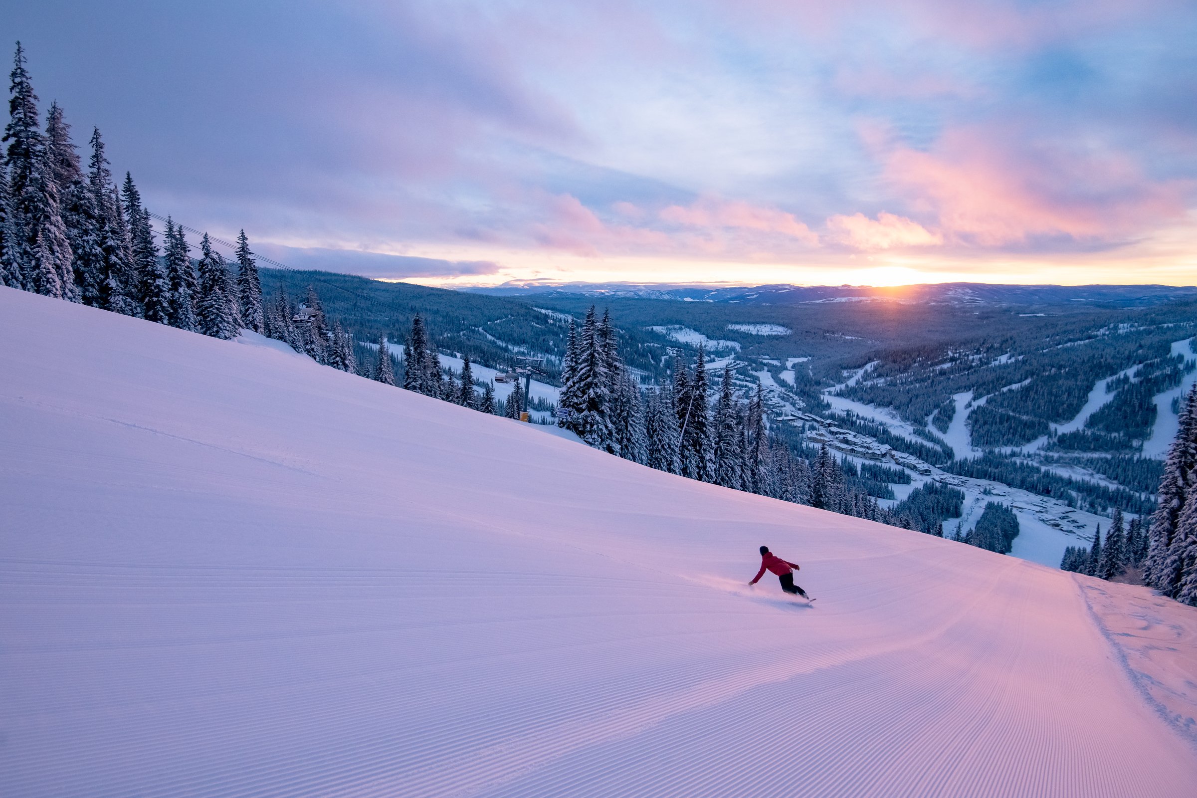 A person skiing downhill on a snow-covered slope during sunset with a scenic view of snow-covered trees and mountains in the background.