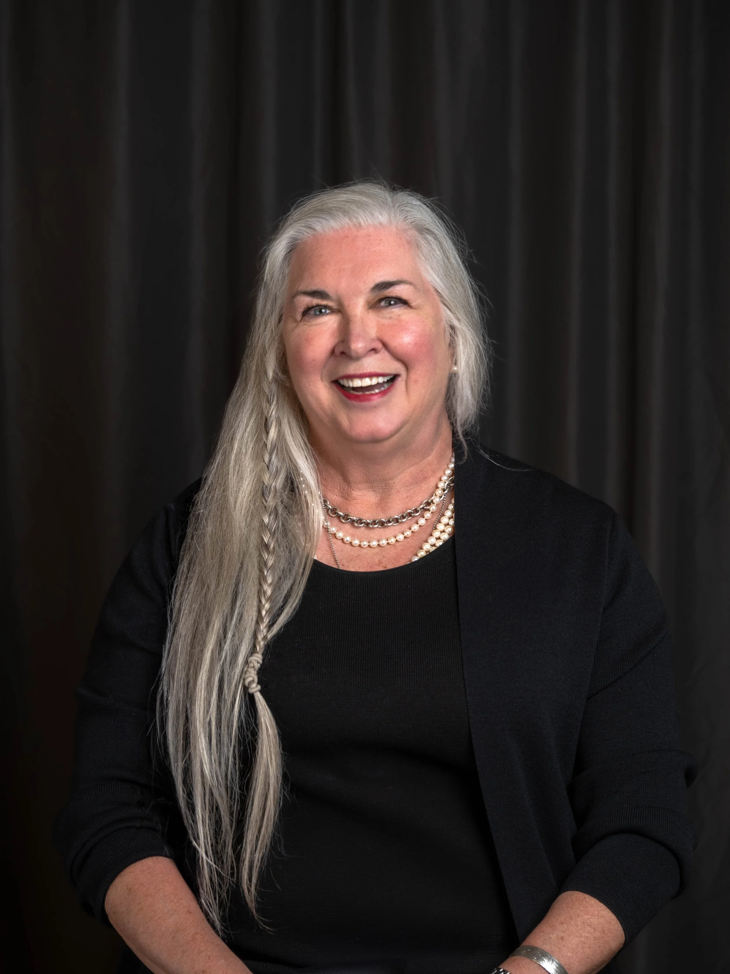 A smiling woman with long gray hair, wearing a black top and blazer, layered pearl and chain necklaces, and a silver bracelet, posing in front of a dark curtain.