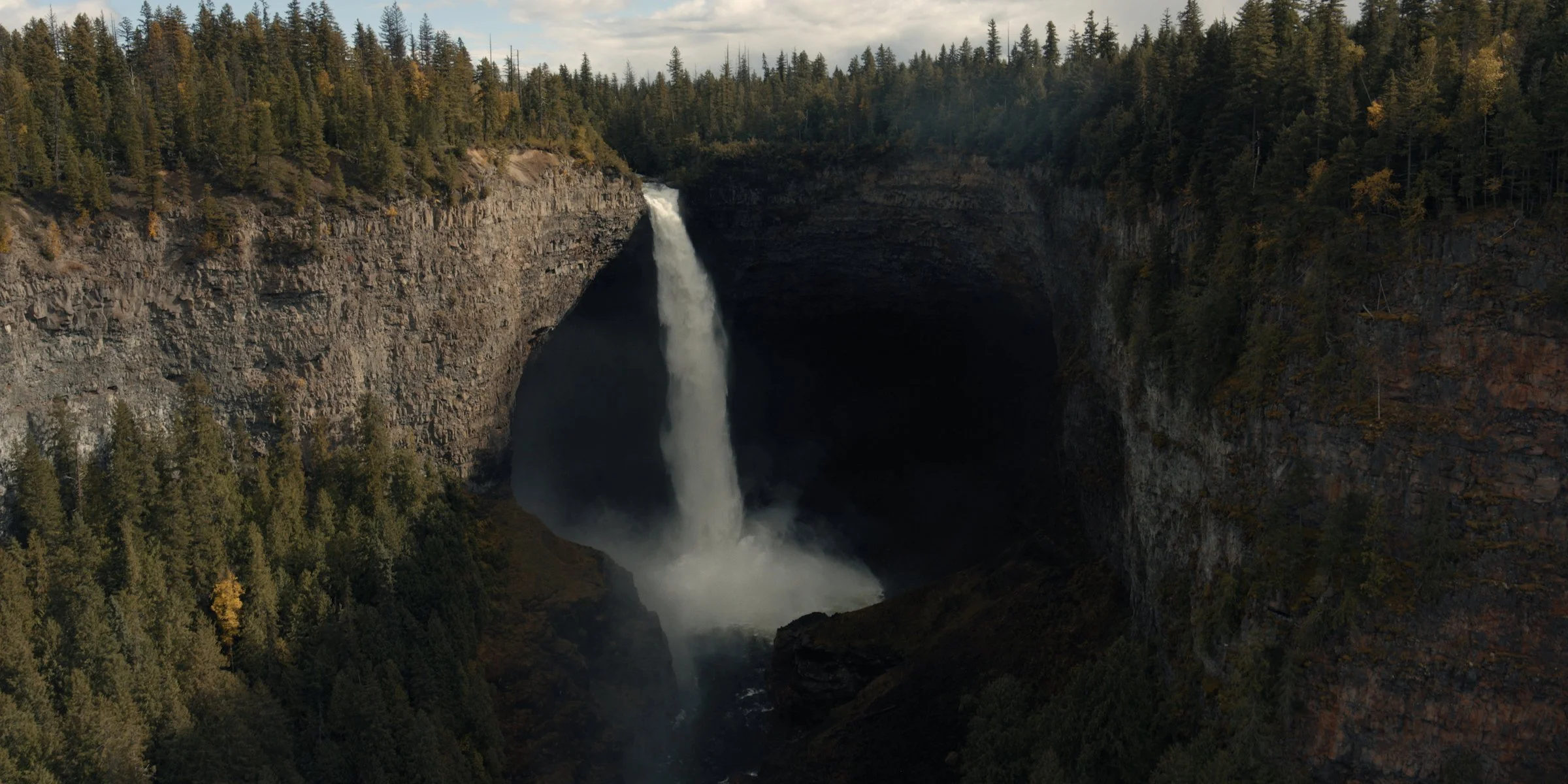 A waterfall flowing into a deep canyon surrounded by dense forest with autumn foliage.