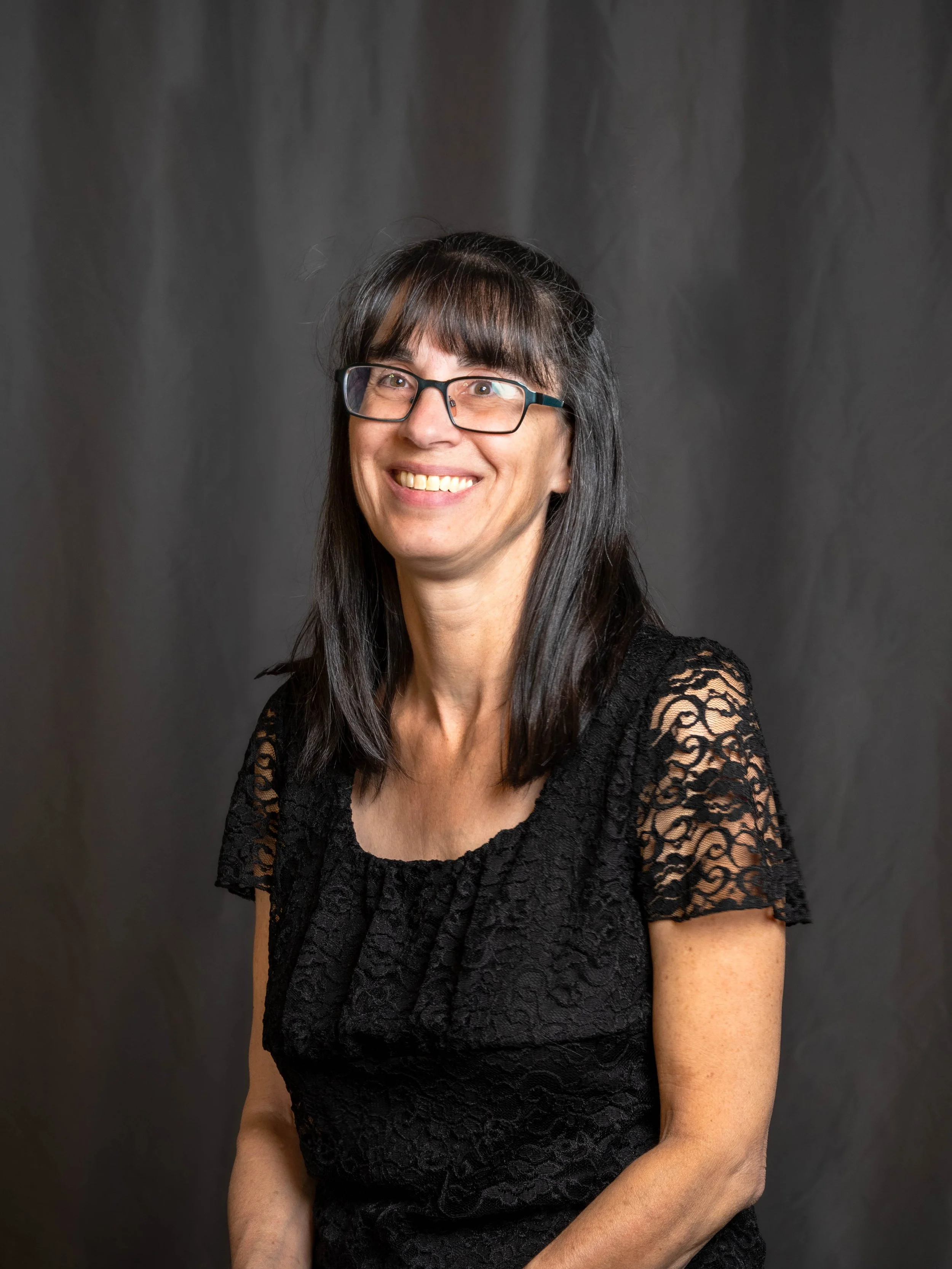 A woman with glasses, long dark hair with bangs, smiling, wearing a black lace top, against a dark backdrop.