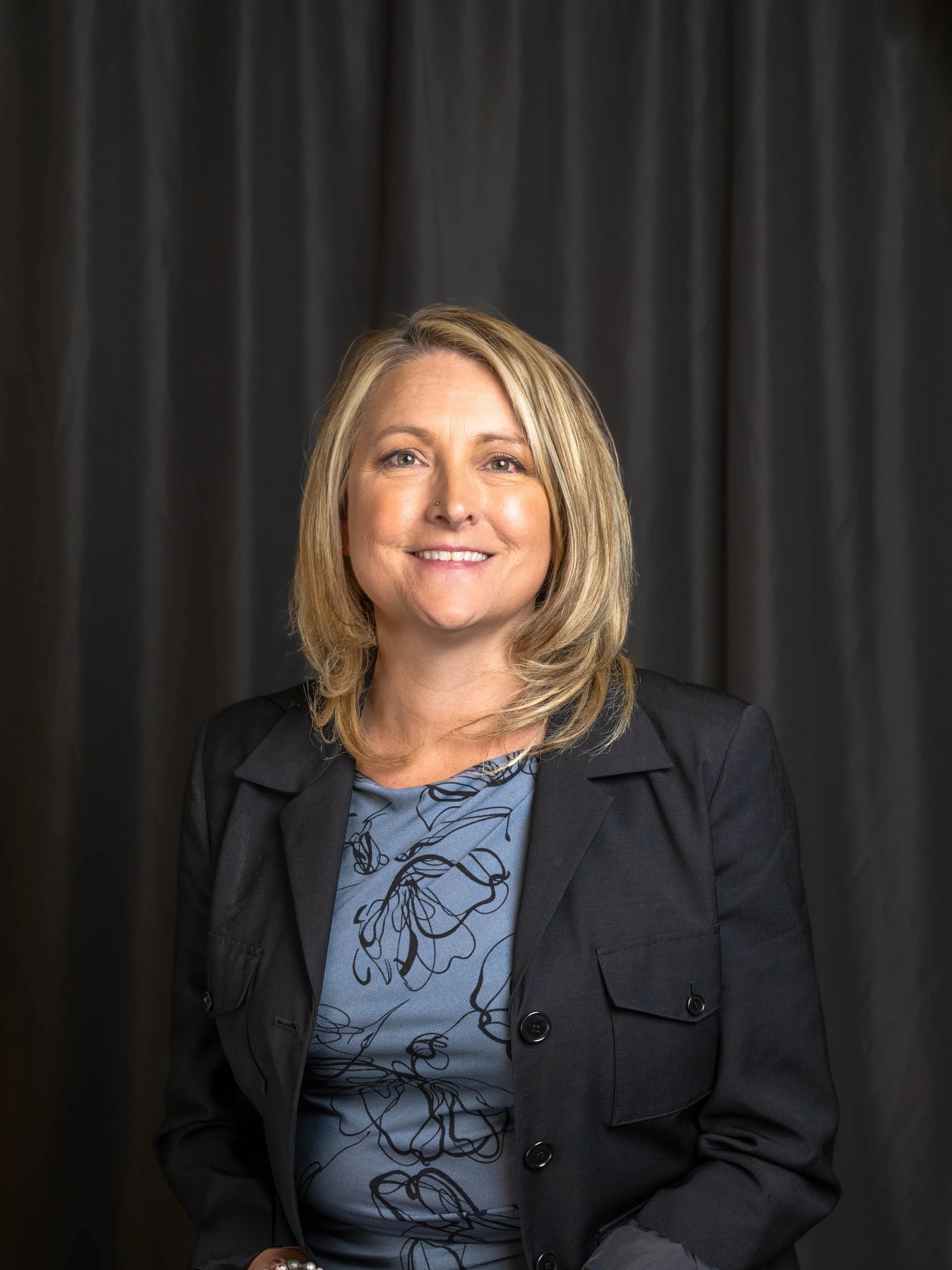 A smiling woman with shoulder-length blonde hair, wearing a black blazer and a blue patterned blouse, standing in front of a dark curtain.
