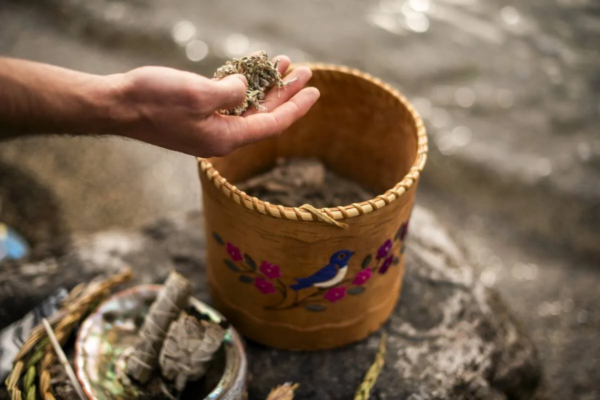 A person holding a handful of dried herbs or plant material over a decorated container with a bird painting, placed on a rocky surface outdoors.