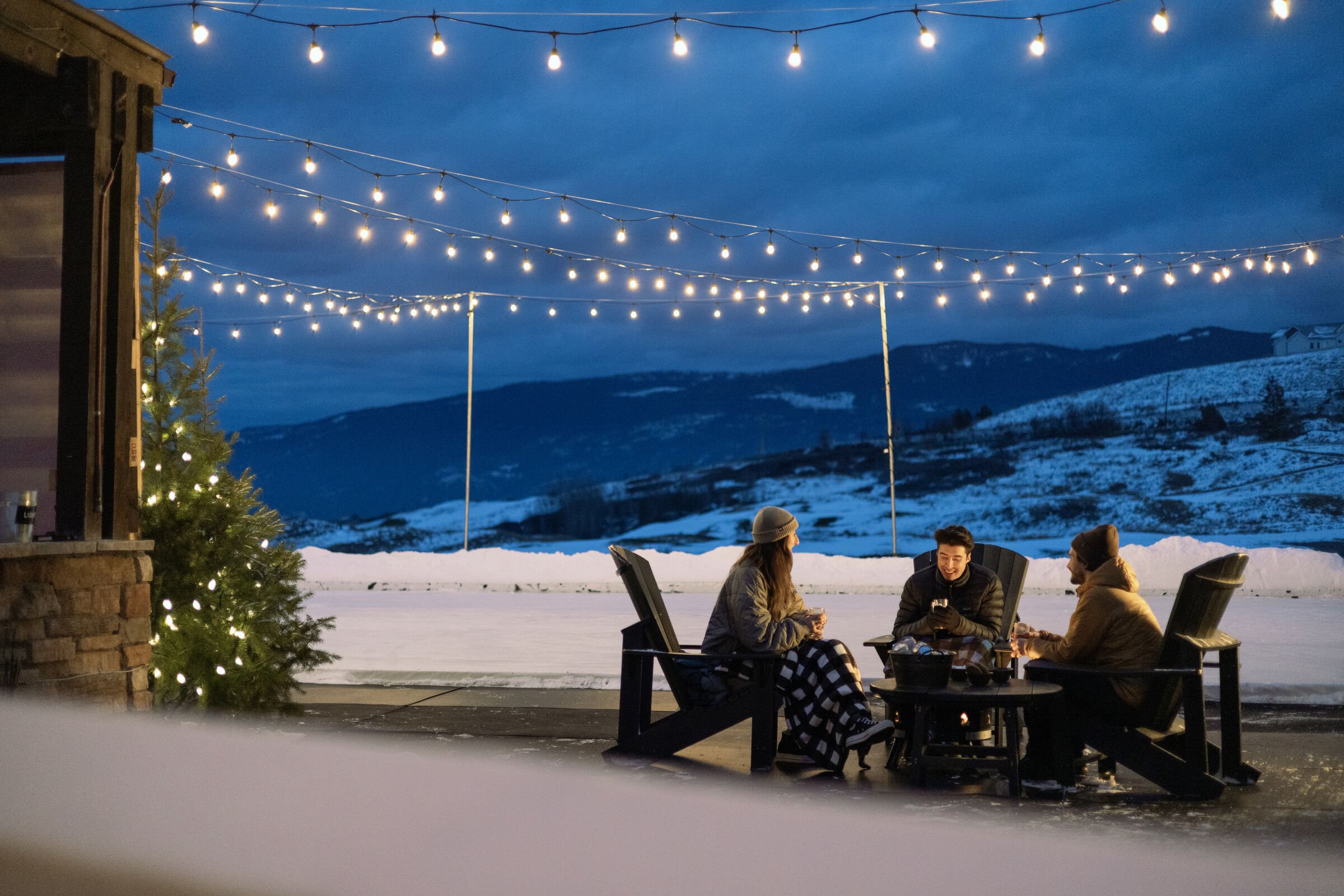 Three people sitting around a fire pit outside during winter evening with snow on the ground, string lights overhead, and in the background a snow-covered mountain landscape.