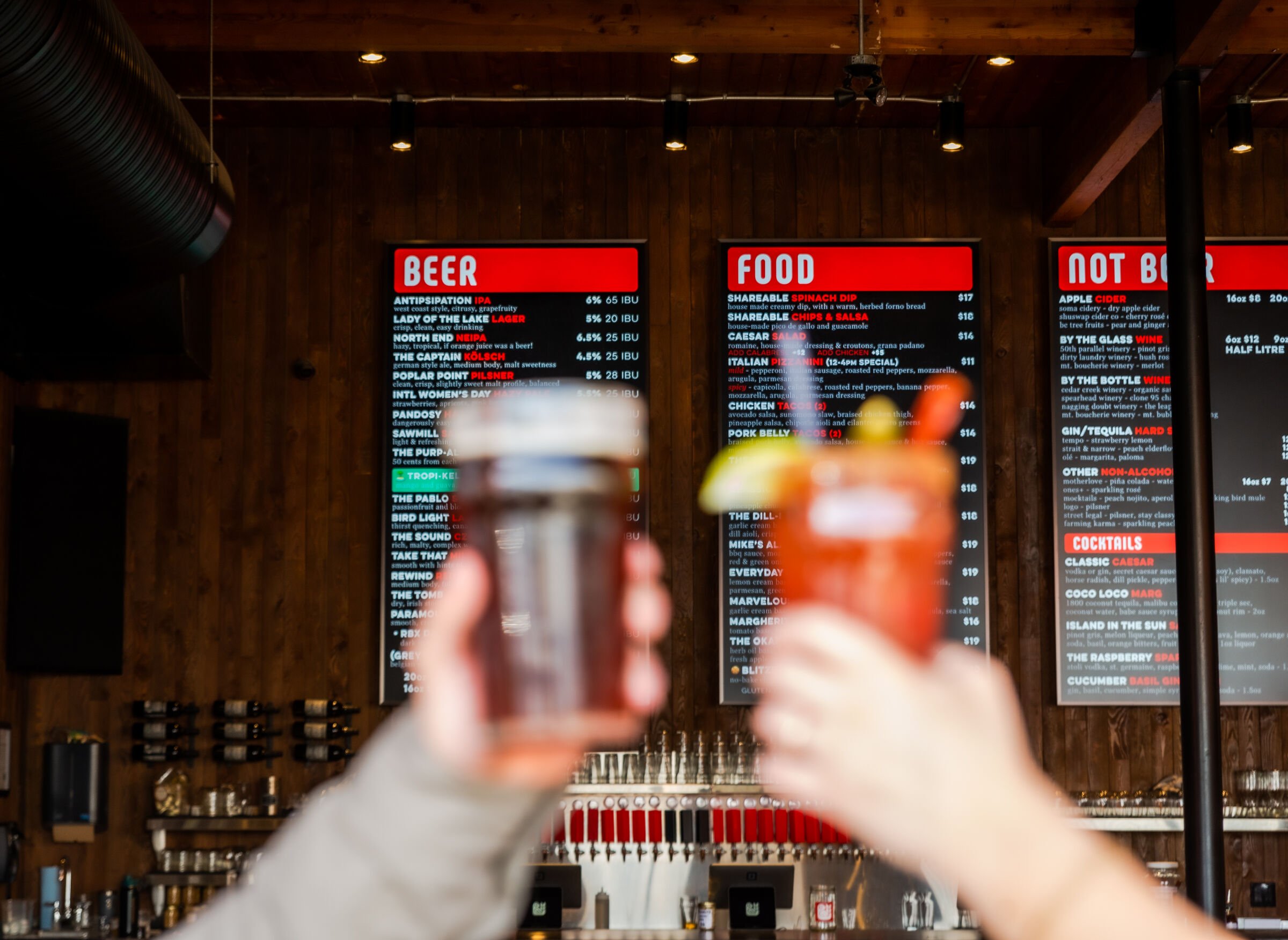 Blurred hand holding a glass of beer in front of digital menu boards at a bar, with various beer, food, and cocktail options displayed.