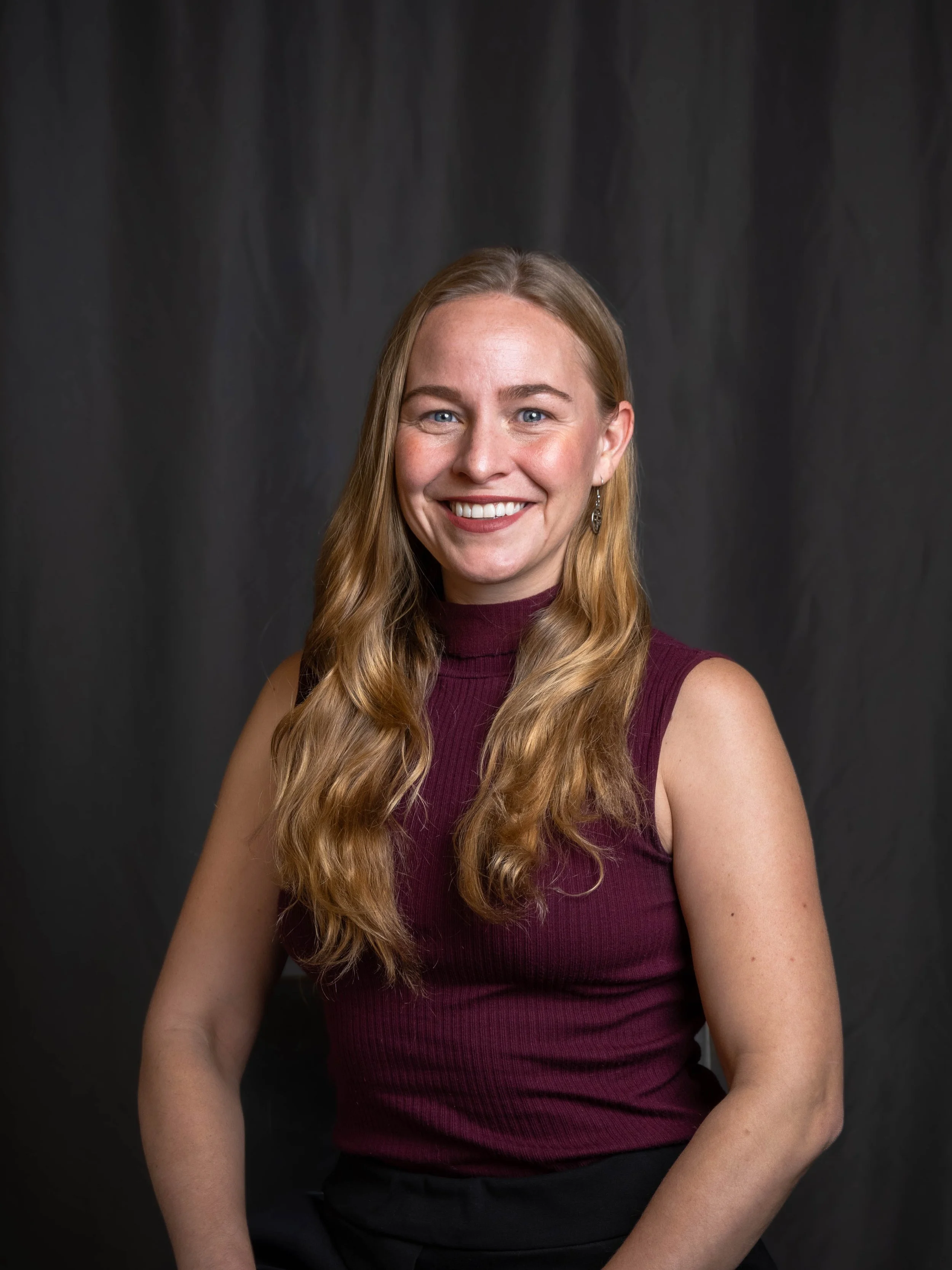 A woman with long wavy blonde hair, blue eyes, and a fair complexion, smiling in front of a dark backdrop. She is wearing a sleeveless turtleneck top in burgundy and earrings.