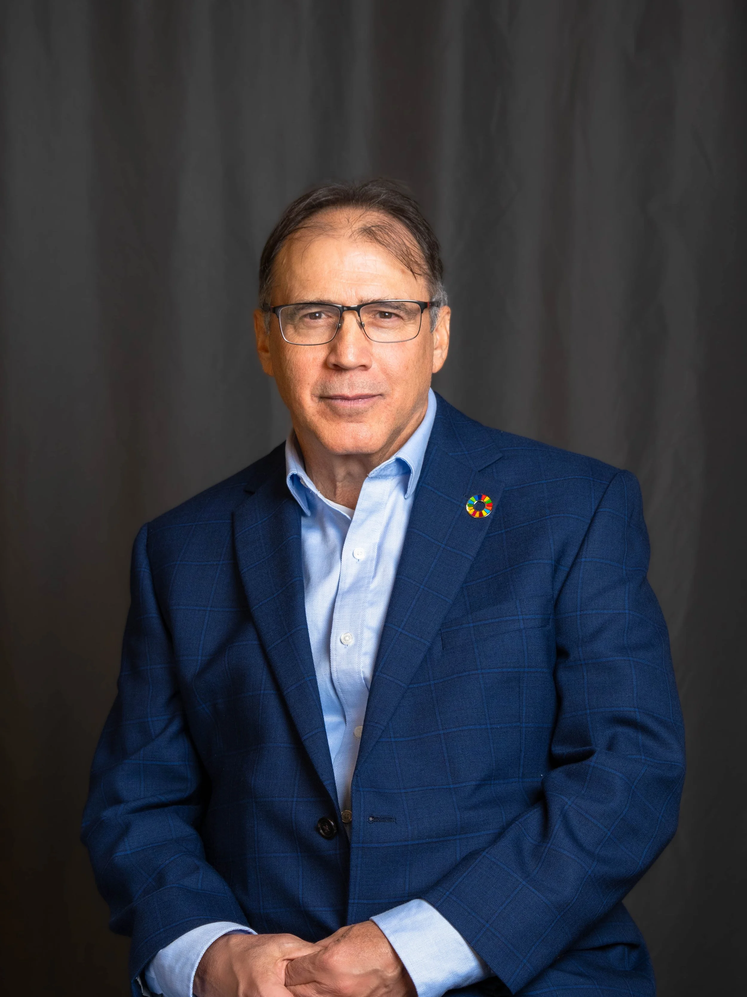 A man wearing glasses and a blue checked suit with a rainbow pin on the lapel, posing against a dark gray backdrop.