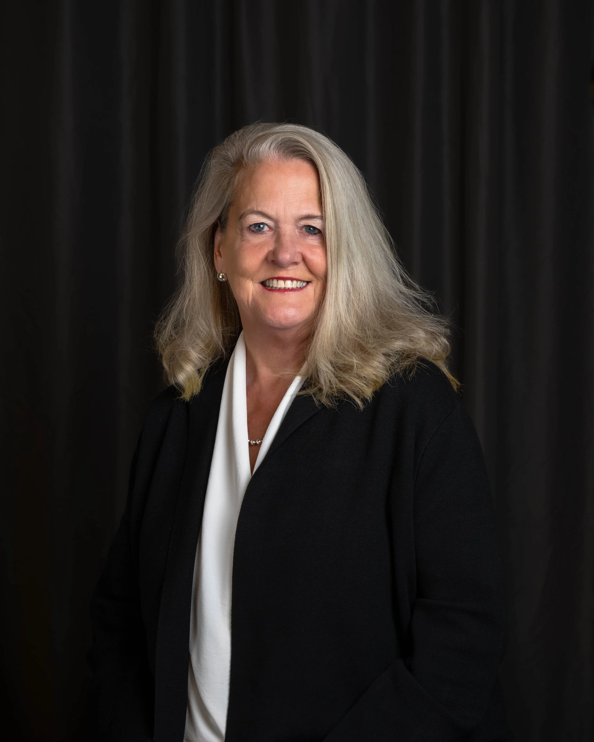 Professional woman with shoulder-length gray hair, wearing a black blazer over a white blouse, smiling against a black curtain background.