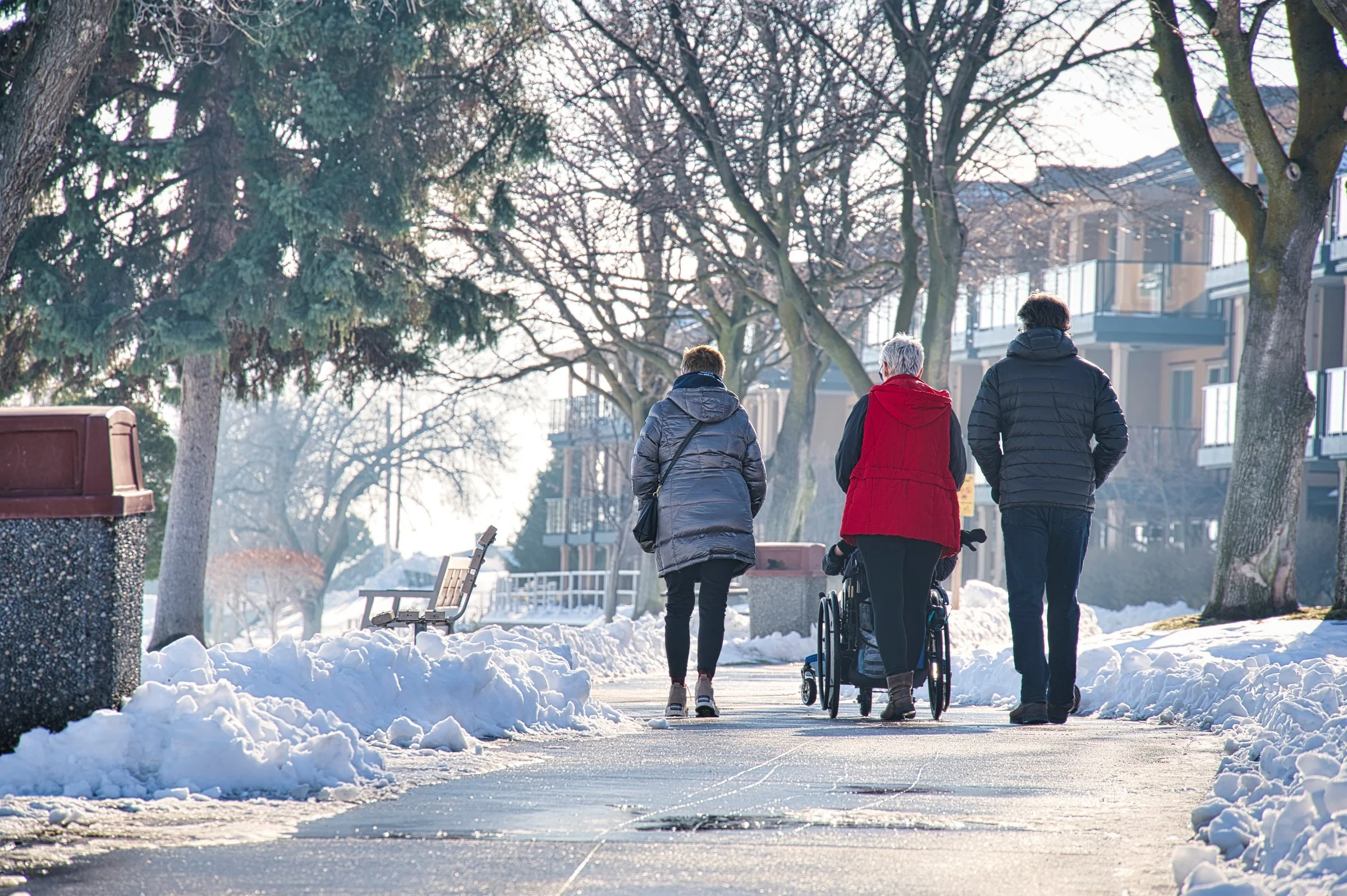 Three people walking on a snow-covered sidewalk in a residential neighborhood with leafless trees and apartment buildings during winter.
