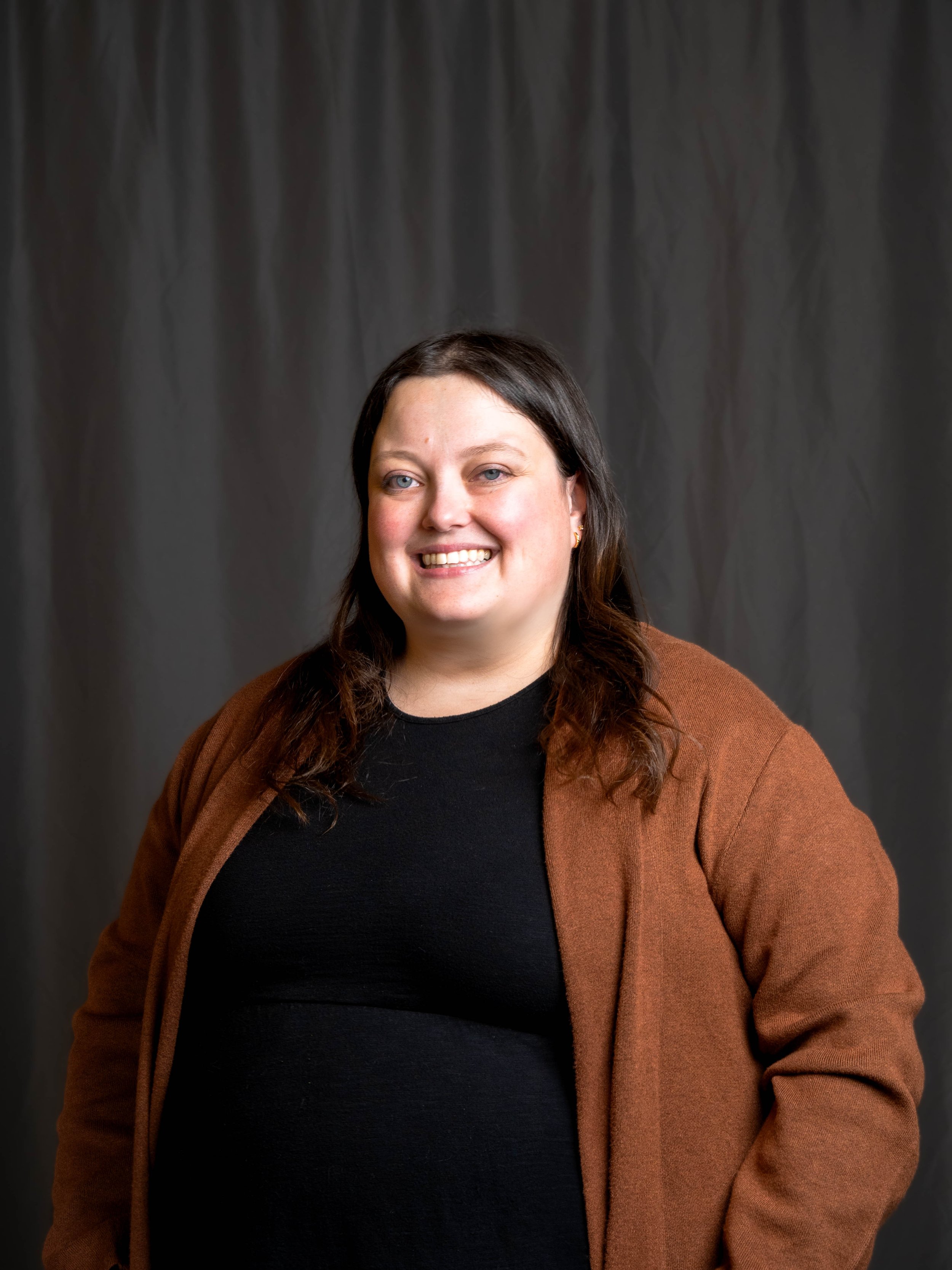 A woman with long brown hair, wearing a black top and a brown jacket, smiling at the camera, standing against a gray backdrop.