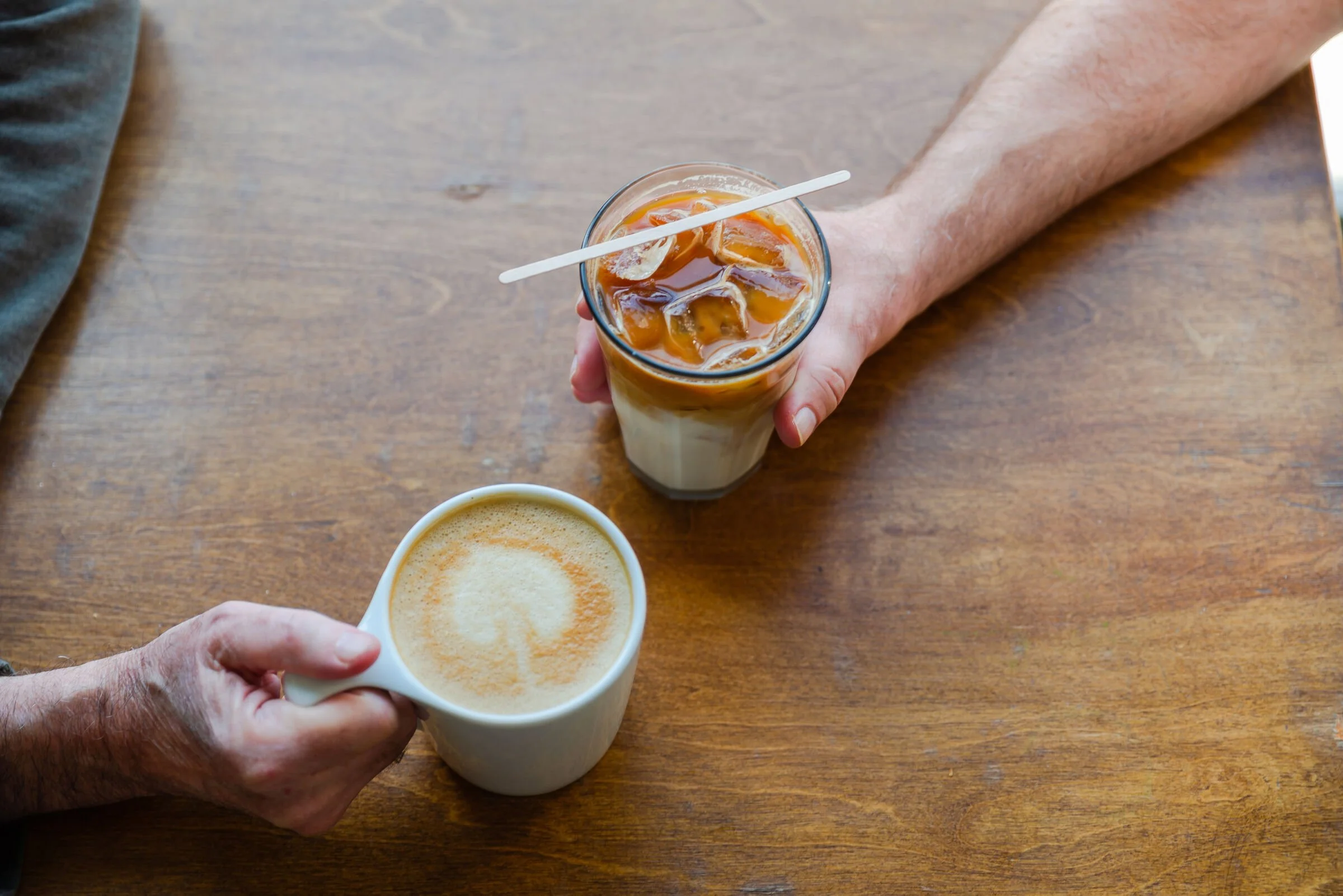 A person holding a glass of iced coffee with ice cubes and a stir stick, and another person holding a mug of frothy coffee or latte, on a wooden table.