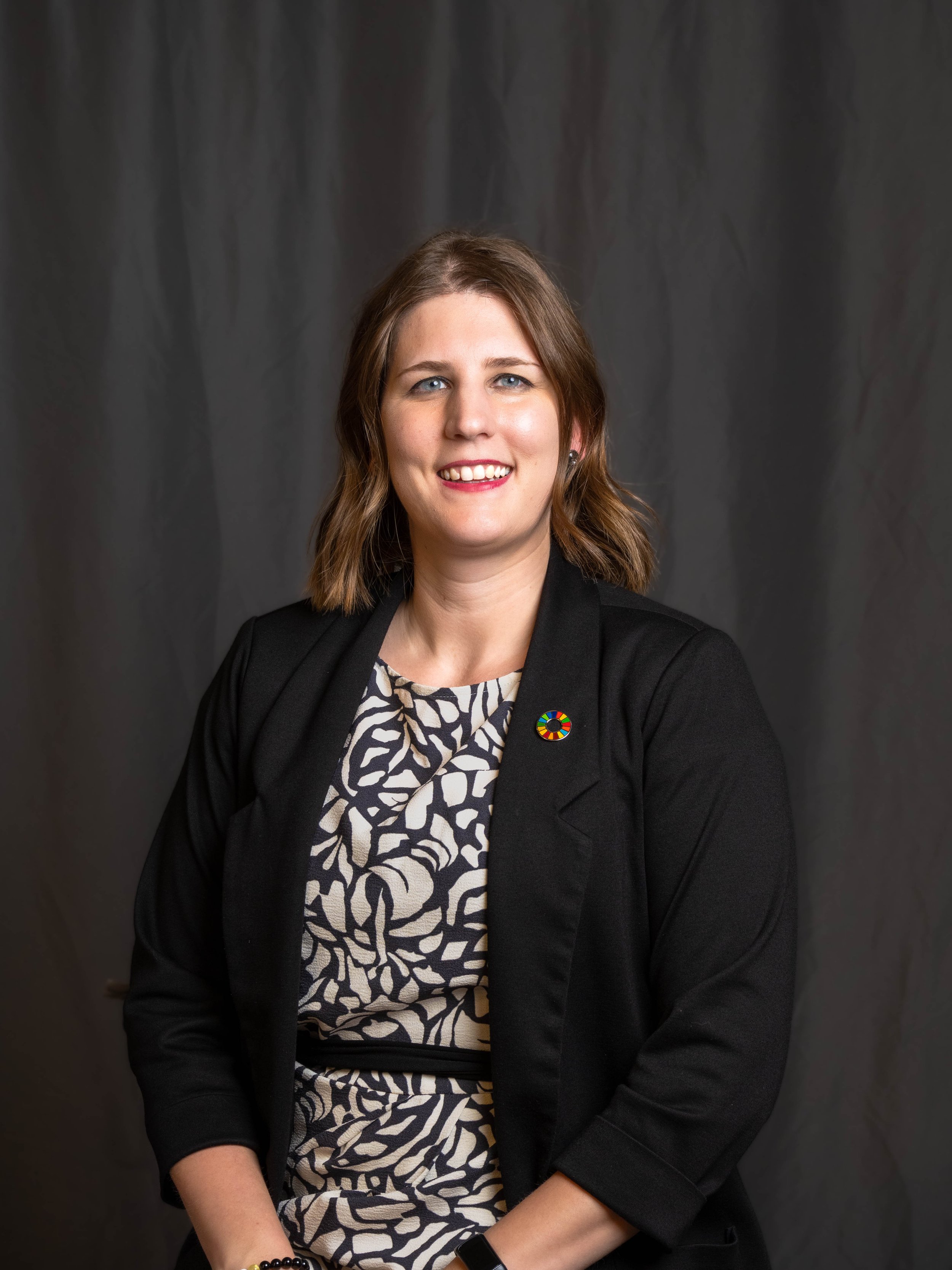 A woman with shoulder-length brown hair, wearing a black blazer with a multicolored circular pin, and a patterned dress, seated in front of a dark gray background, smiling at the camera.