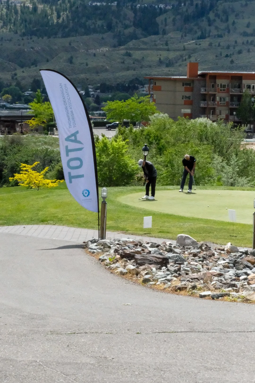 Two people playing golf on a putting green with a flag that reads 'TOTA' and 'Thompson & Morgan Tourism Association', surrounded by trees and rocks, with buildings and mountains in the background.