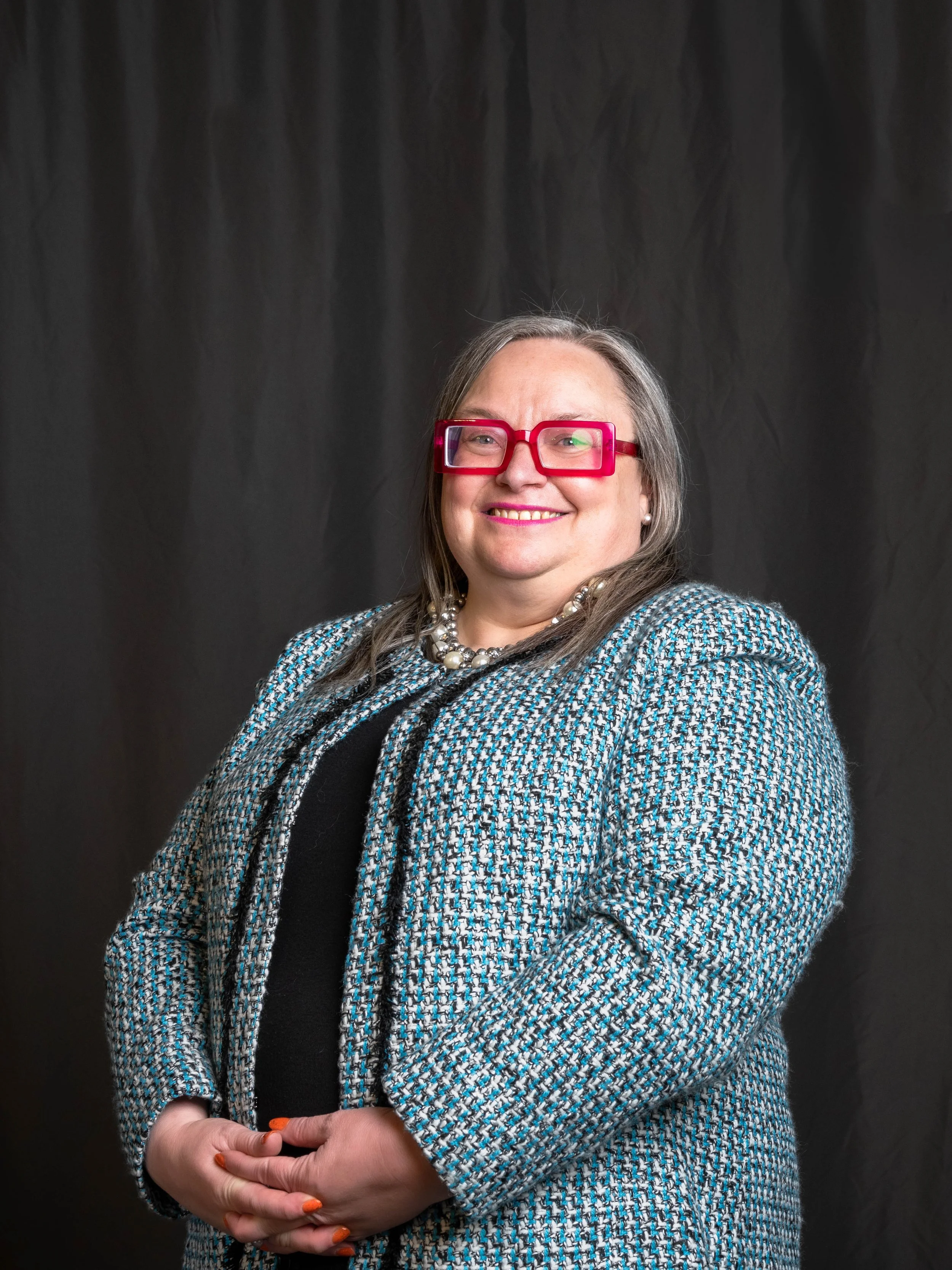 A woman with long gray hair, wearing red glasses, a pearl necklace, a black top, and a patterned blazer, is smiling and standing against a dark curtain background.