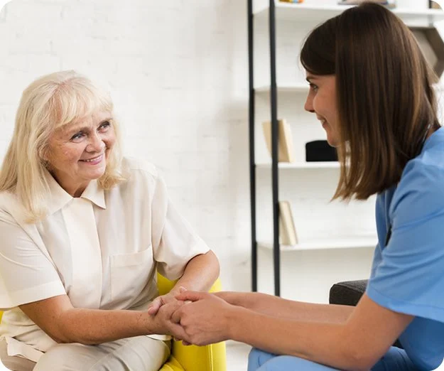 An in-home care nurse in blue scrubs sitting on a couch and holding the hand of an older woman in a bright yellow chair.