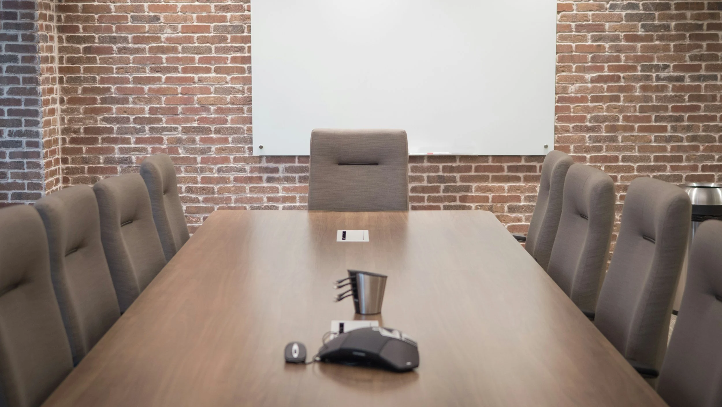 Empty conference room with a long wooden table, beige chairs, a whiteboard, and brick walls.
