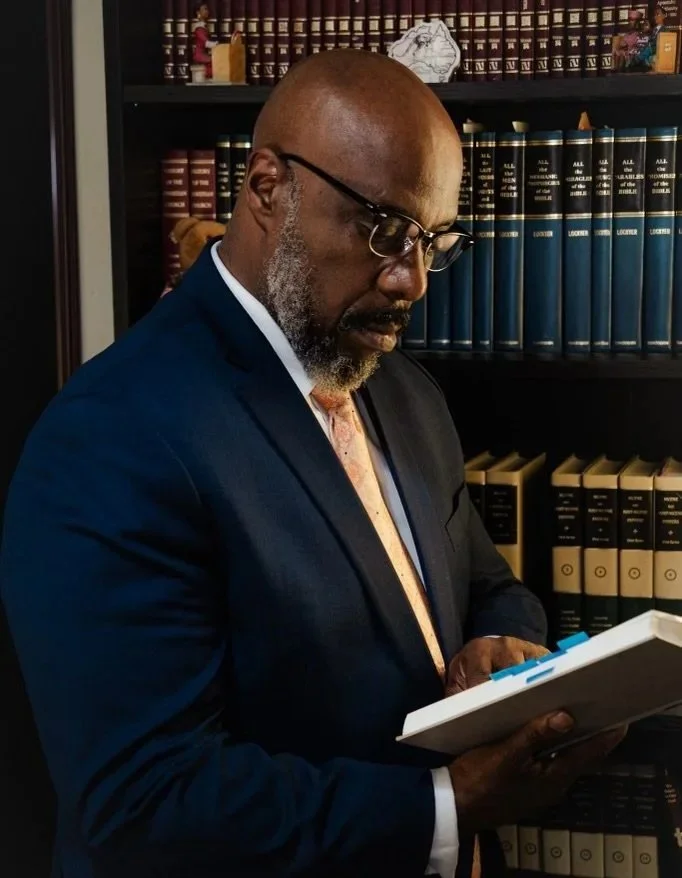 A man in a dark suit, light pink tie, and glasses is reading a document in a library or office, with shelves of law books in the background.